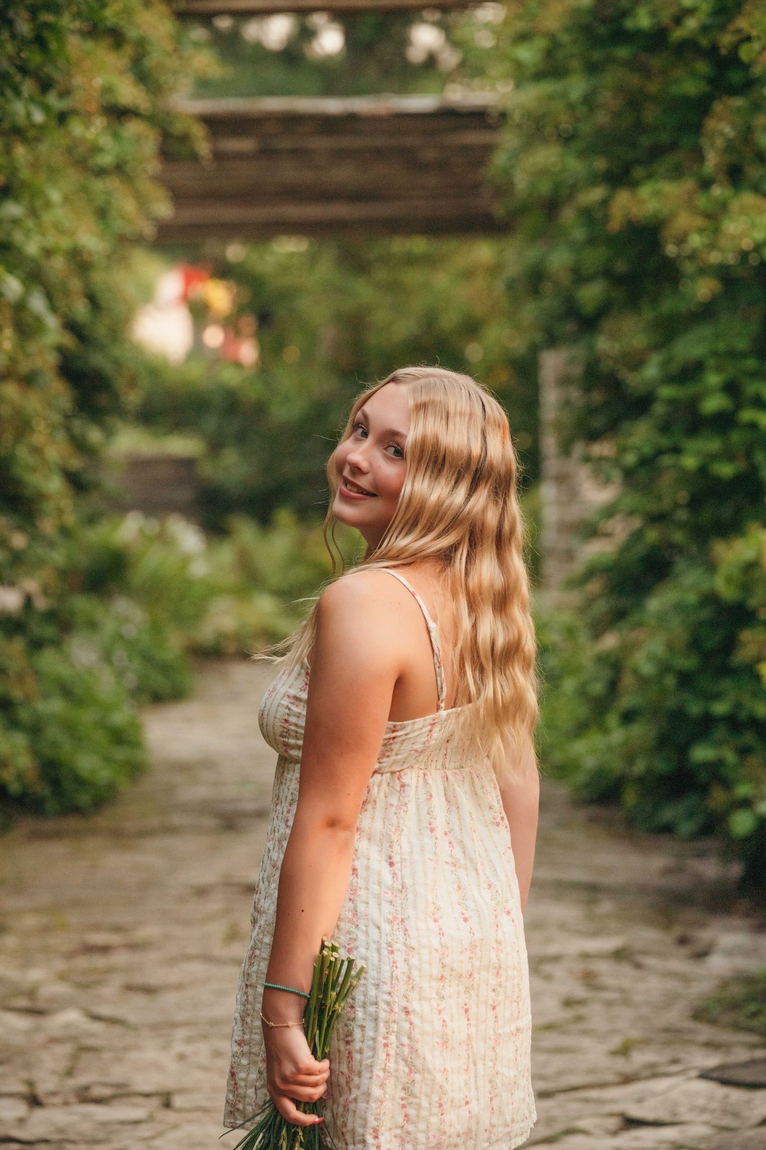 Young blonde woman in floral sundress holding flowers, smiling in a lush green garden pathway.