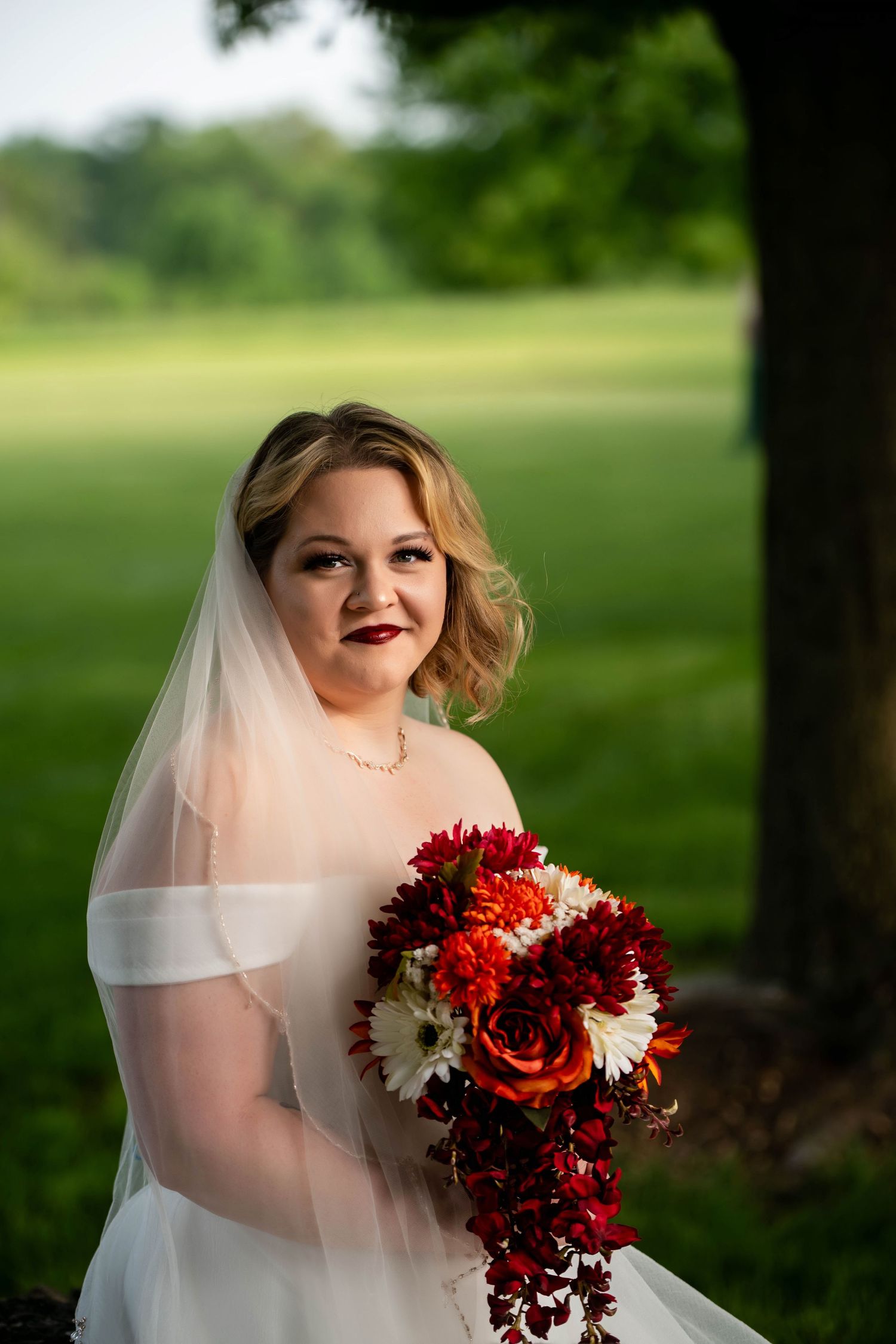 Bride in white dress holds vibrant red and white bouquet outdoors.