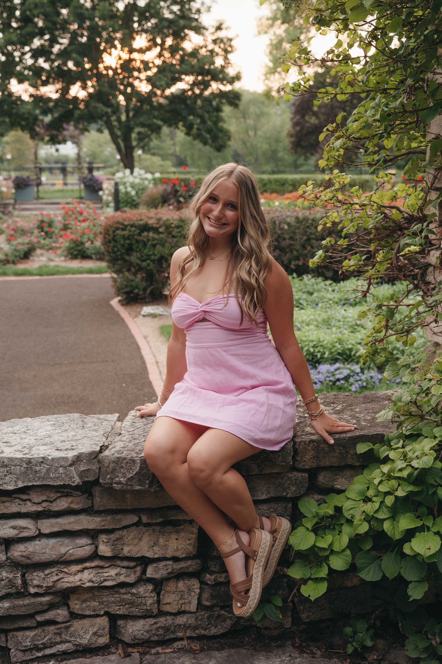 Young woman in pink strapless mini dress sitting on stone wall in lush garden park at sunset.