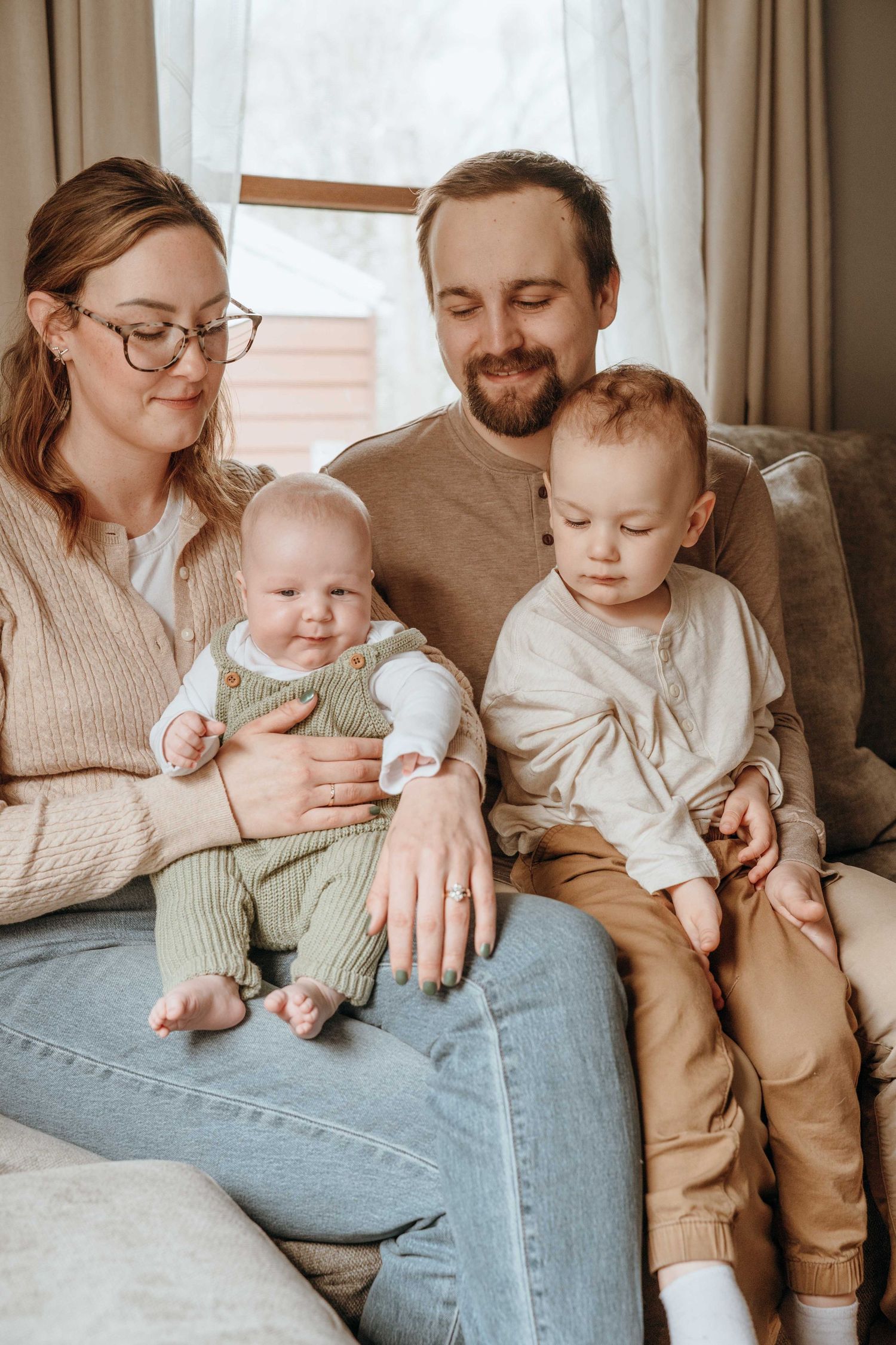 Happy young family with two babies sitting together on a couch in a cozy, warmly lit living room.
