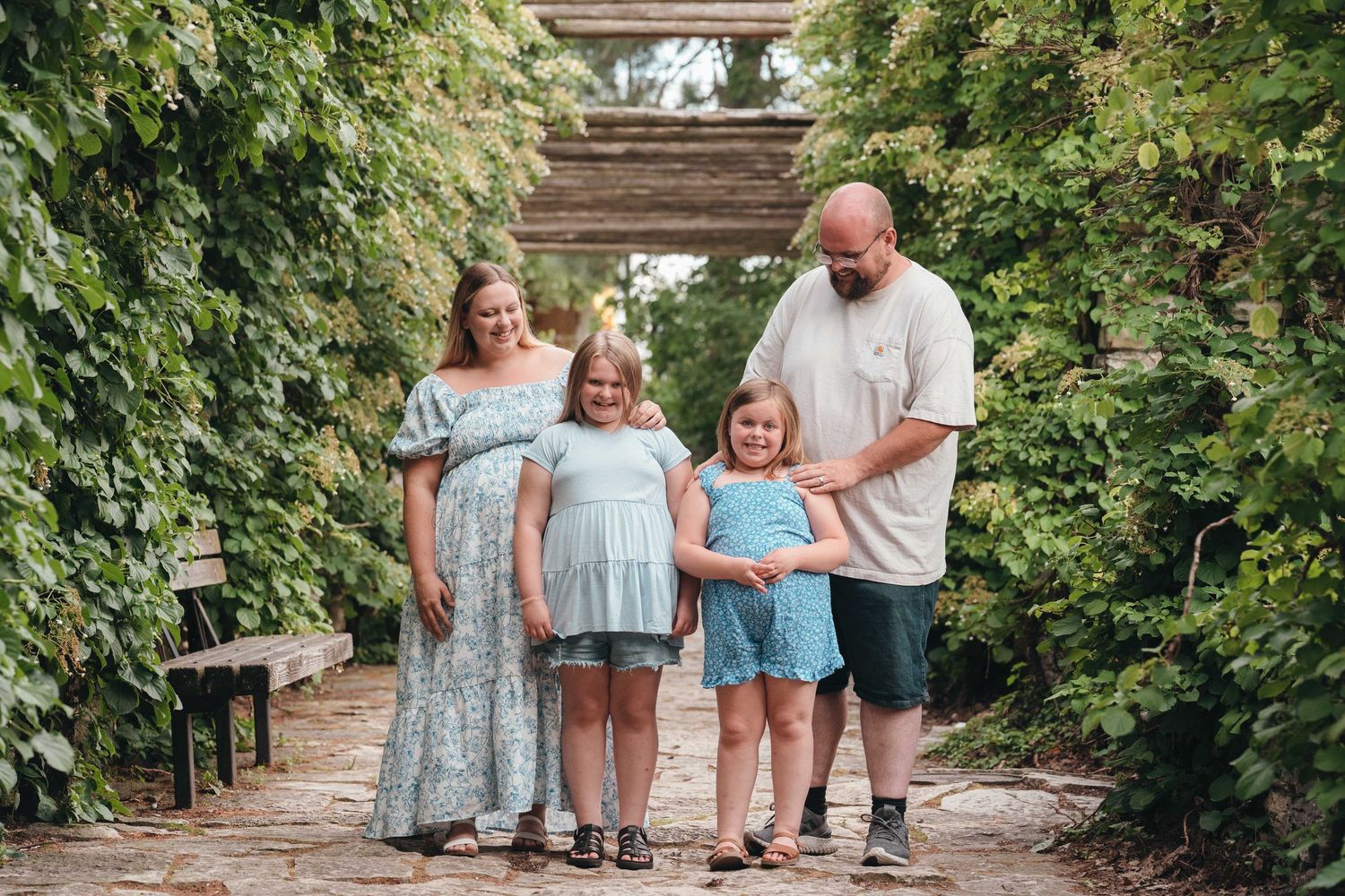 Family of four poses on a garden path, parents with two young girls surrounded by lush green ivy archway.