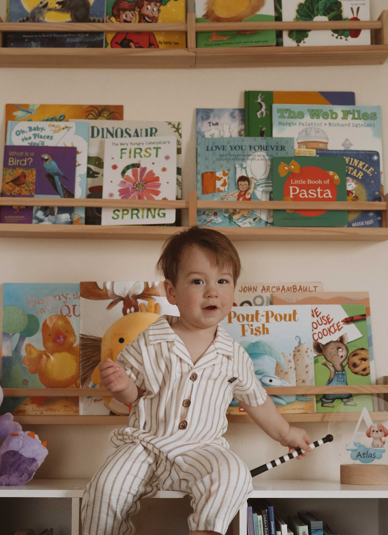 Toddler in striped pajamas sitting on a shelf surrounded by colorful children's books including Dinosaur Spring and Pout-Pout Fish.