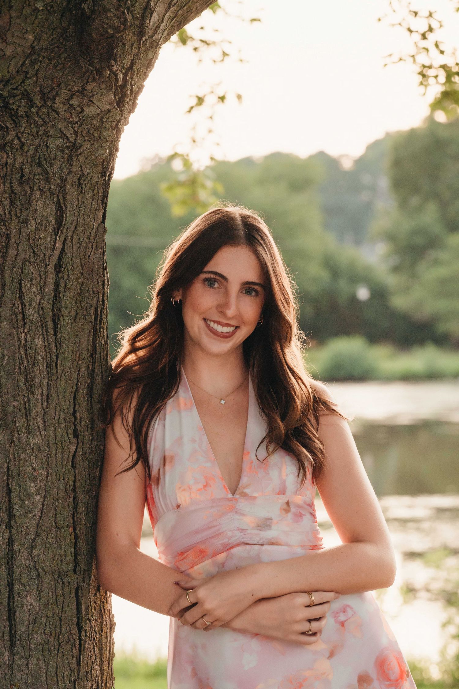 Young woman in floral dress leaning against tree by a lake, smiling outdoors in golden hour light.