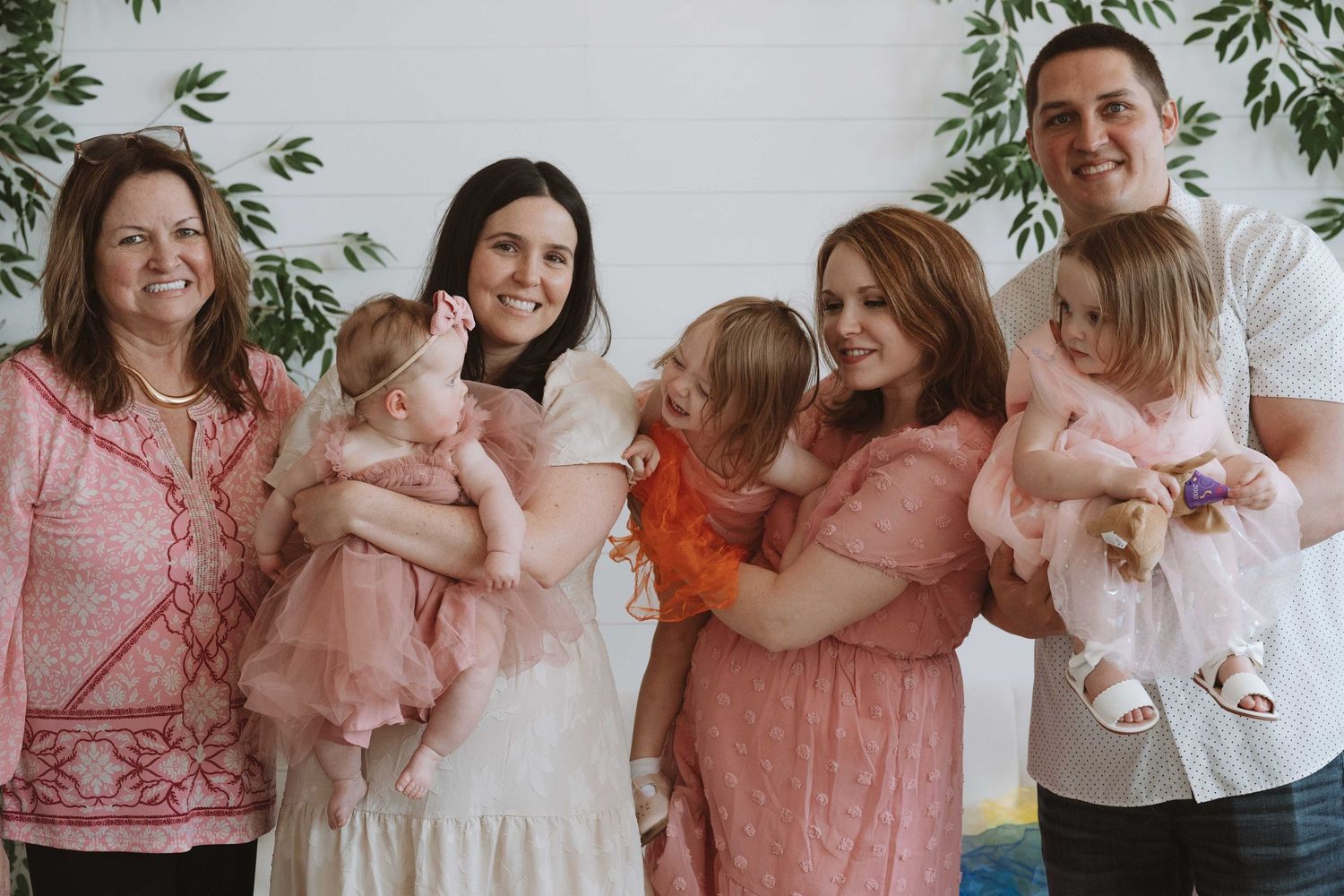 Family group photo with adults holding toddlers in pink outfits, smiling together indoors with greenery backdrop.
