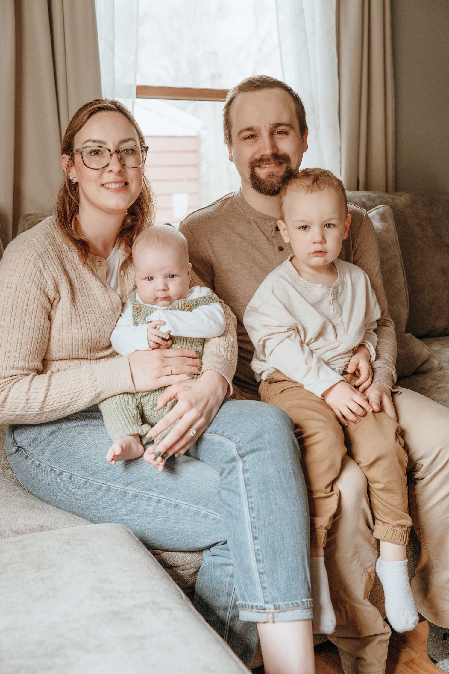A young family of four poses on a couch, with a mother holding a baby and a father holding a toddler, smiling warmly.