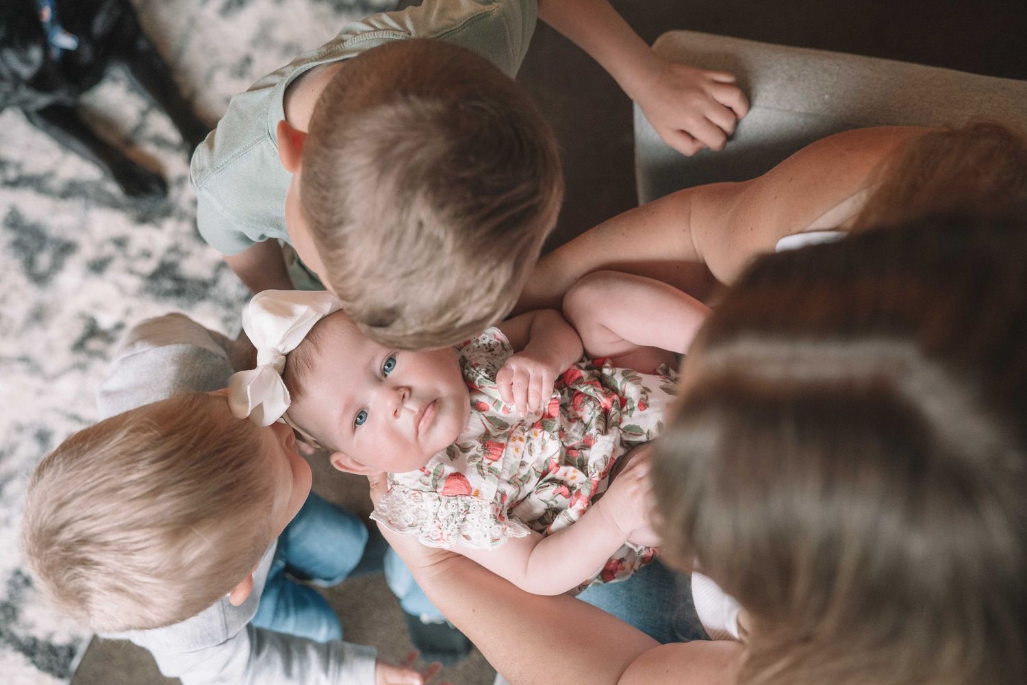 Two young children lovingly kiss and cuddle a smiling baby in a floral outfit on a cozy floor.