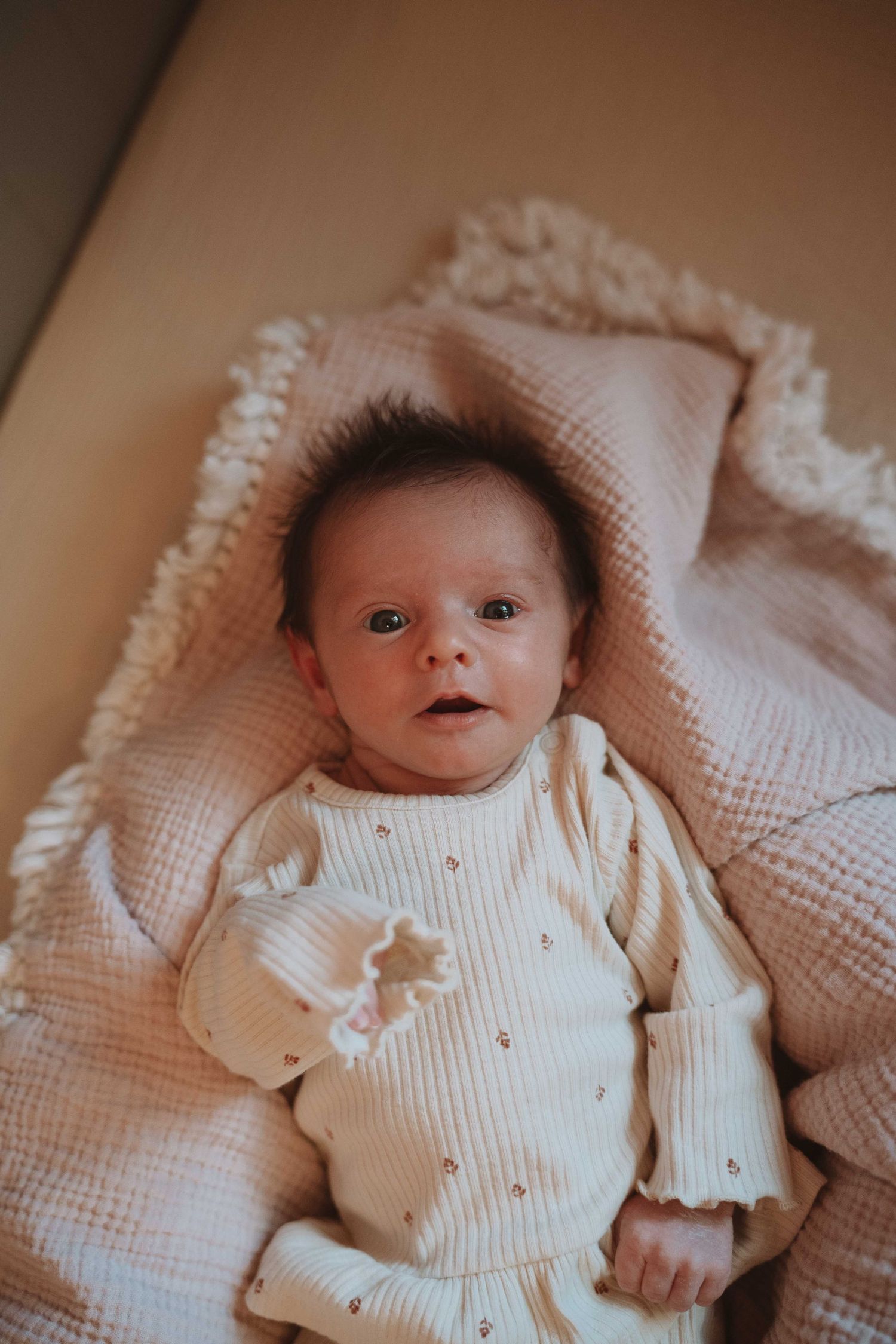 Newborn baby lying on a pink knit blanket wearing a cream ribbed outfit, looking up with wide eyes.