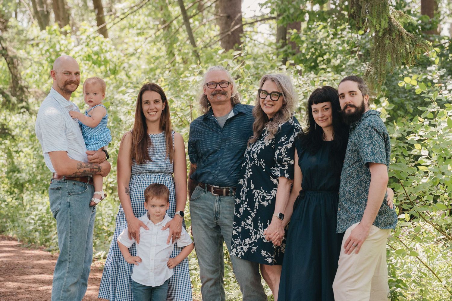 Multi-generational family of seven posing together outdoors in a lush green forest setting for a portrait.