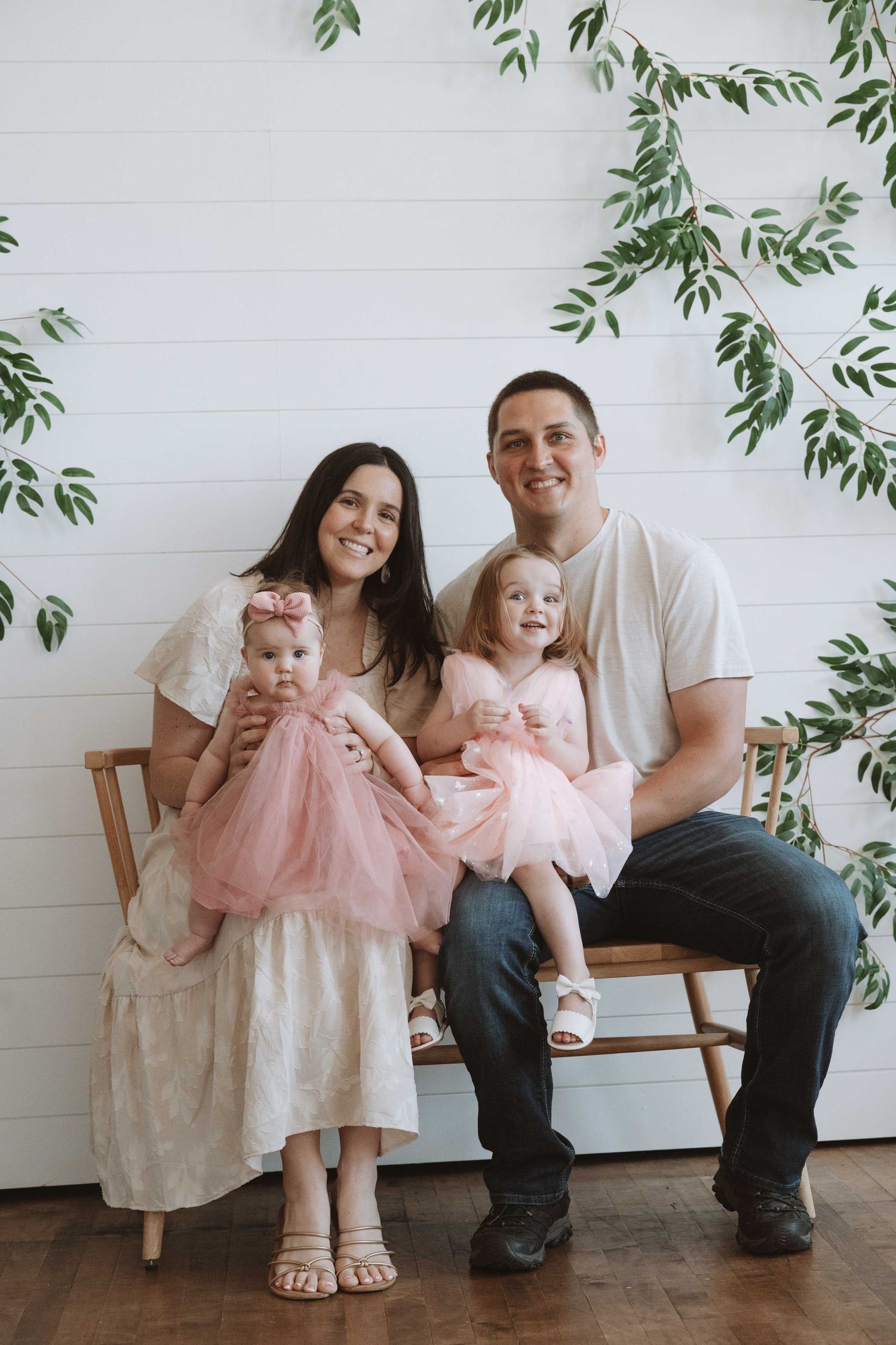 Happy family of four posing for portrait with two young daughters in pink tutus against white shiplap wall with greenery.