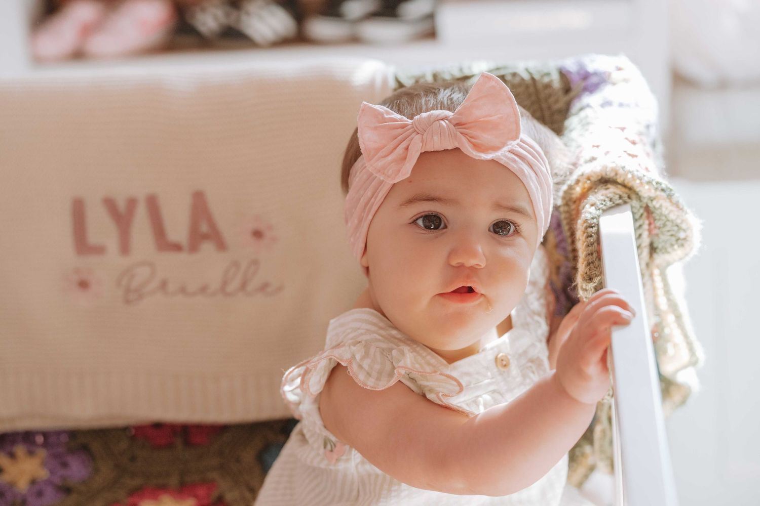 Cute baby girl wearing a pink bow headband sits in a crib next to a personalized blanket reading Lyla Brielle.