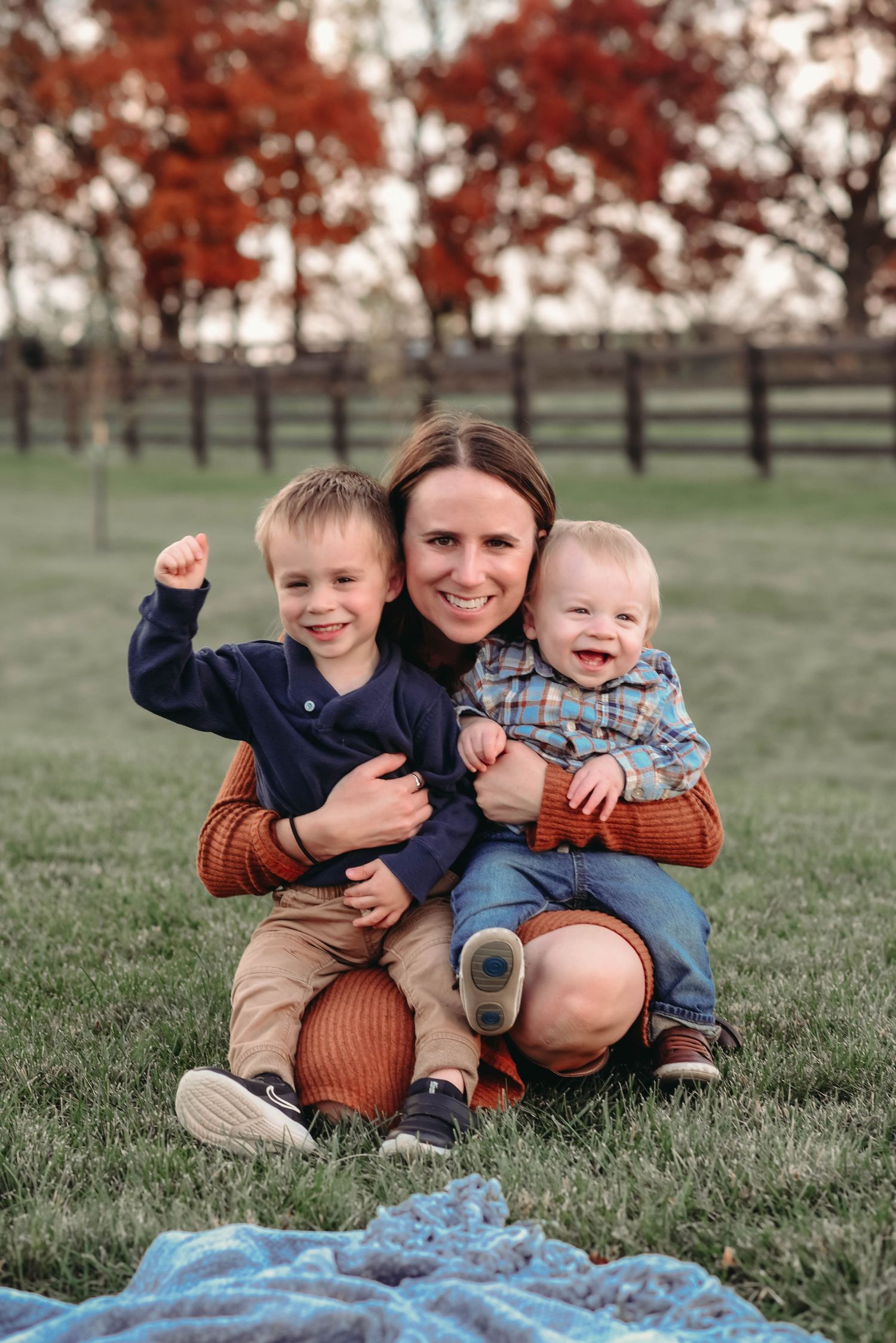 A family sits together on a blanket in a grassy field with red autumn trees in the background.