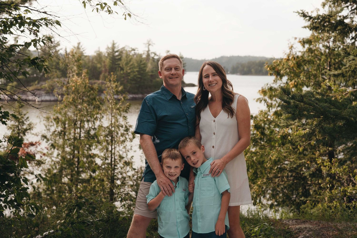 Happy family of four posing outdoors near a lake surrounded by lush green trees in a warm summer setting.