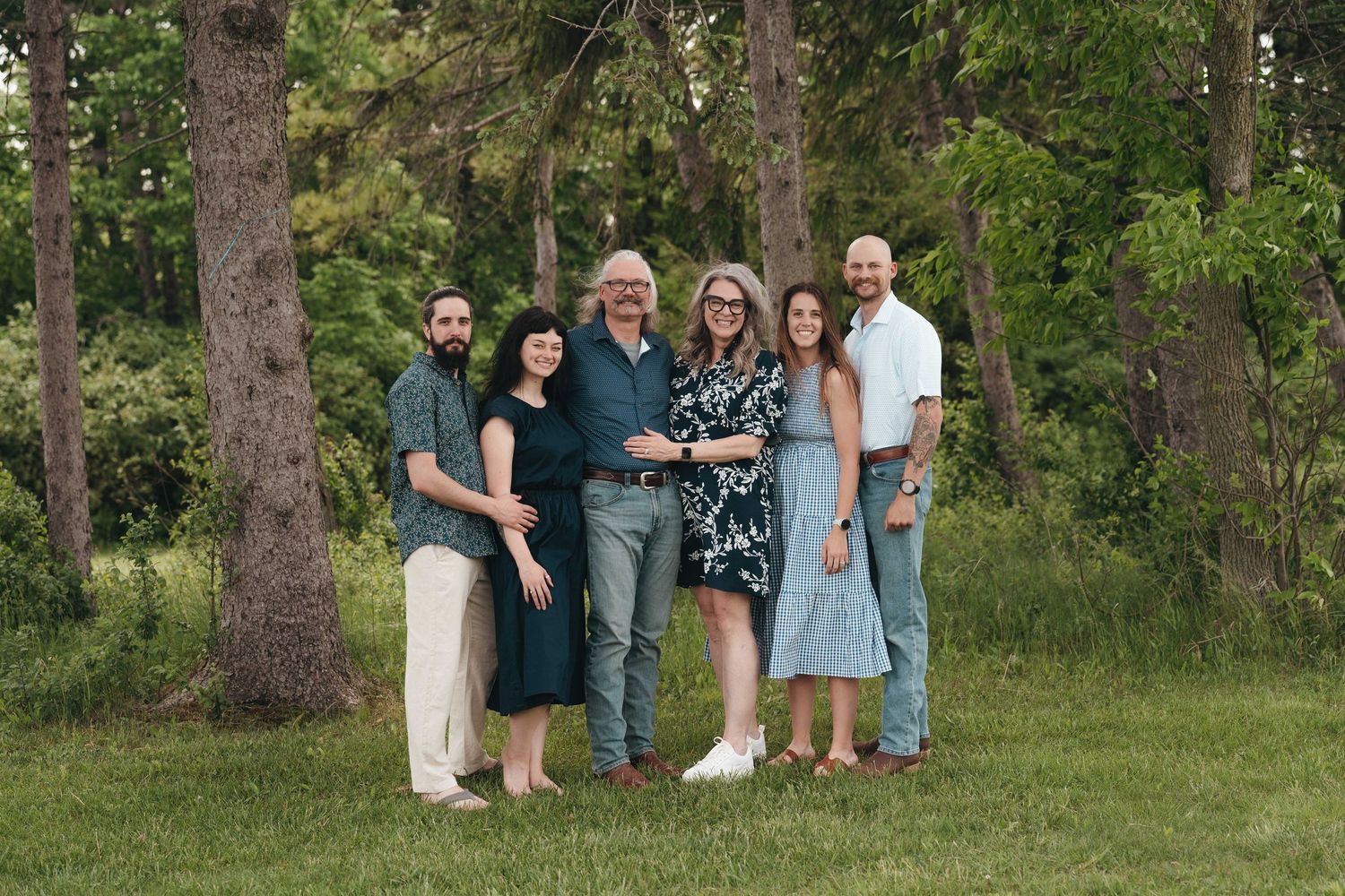 Family of six smiling together outdoors among tall trees on a green lawn during a sunny day.