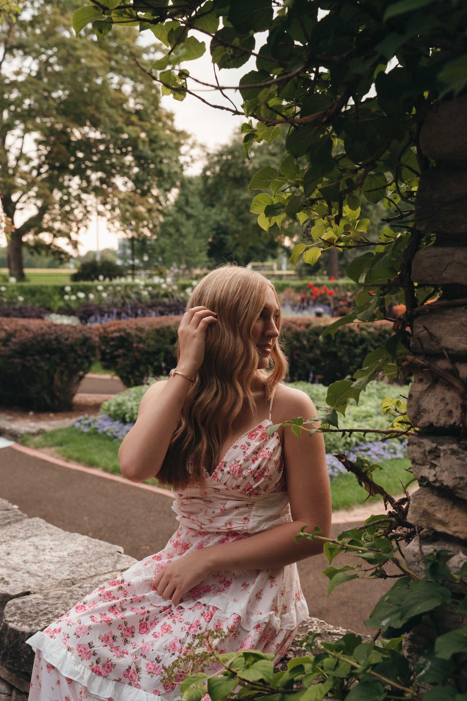 Young woman in floral dress sitting on stone wall in lush garden, looking away with hair blowing gently.