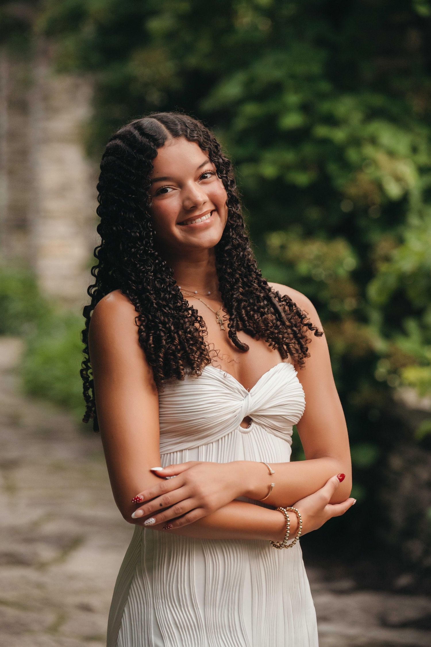 Young woman with curly hair smiling in a white dress, posing outdoors with lush green foliage background.