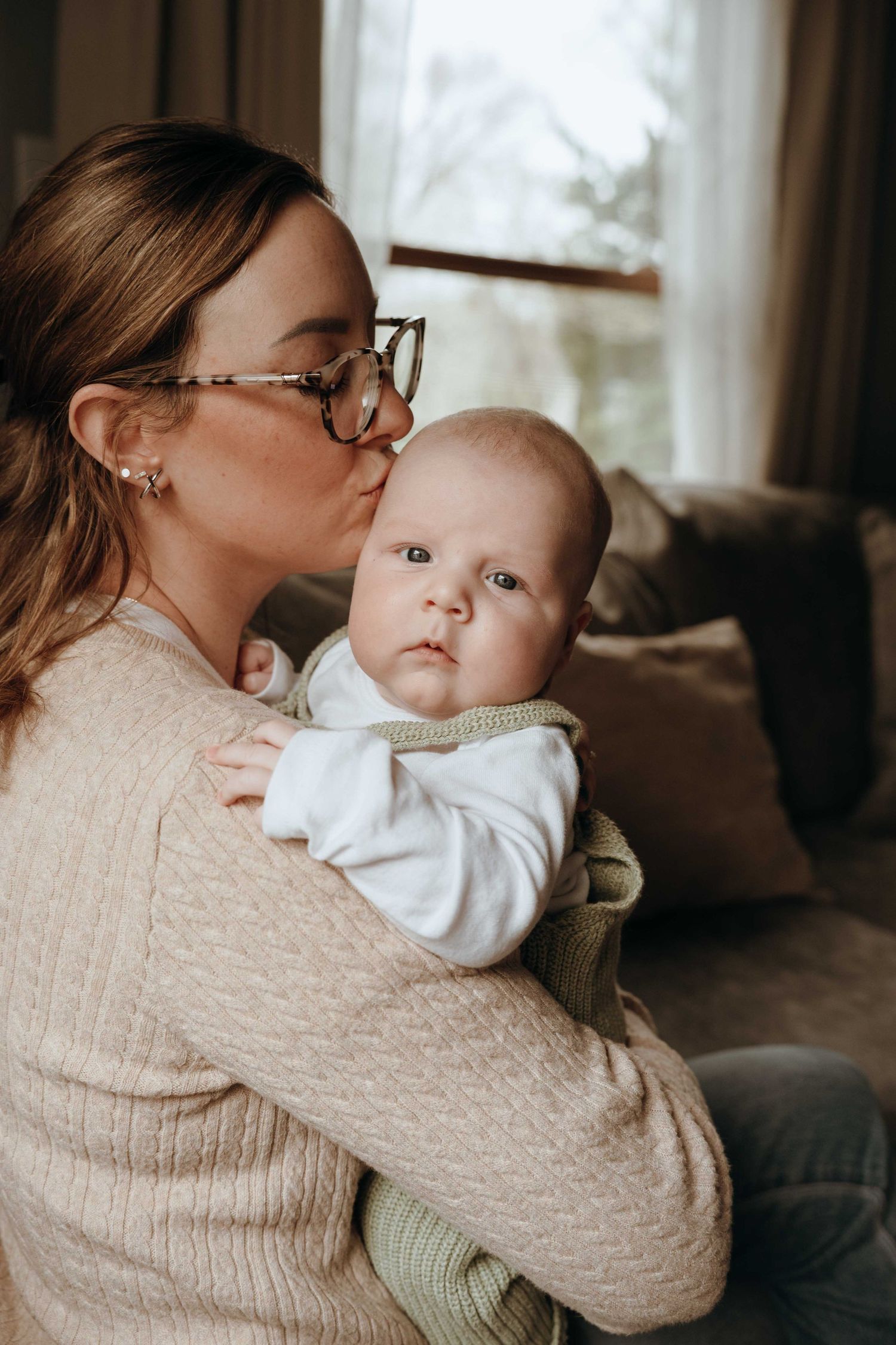 A mother with glasses kisses her newborn baby while holding them in a cozy living room.
