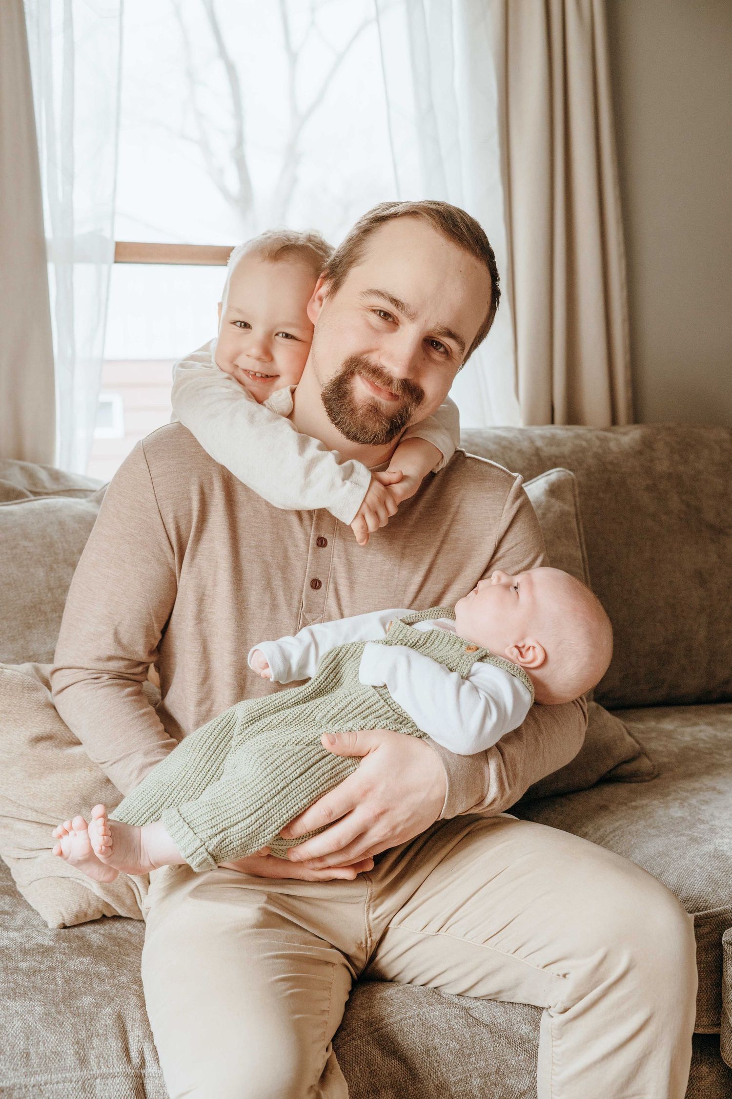 A bearded father sits on a couch holding a newborn baby while a toddler hugs him from behind, smiling.