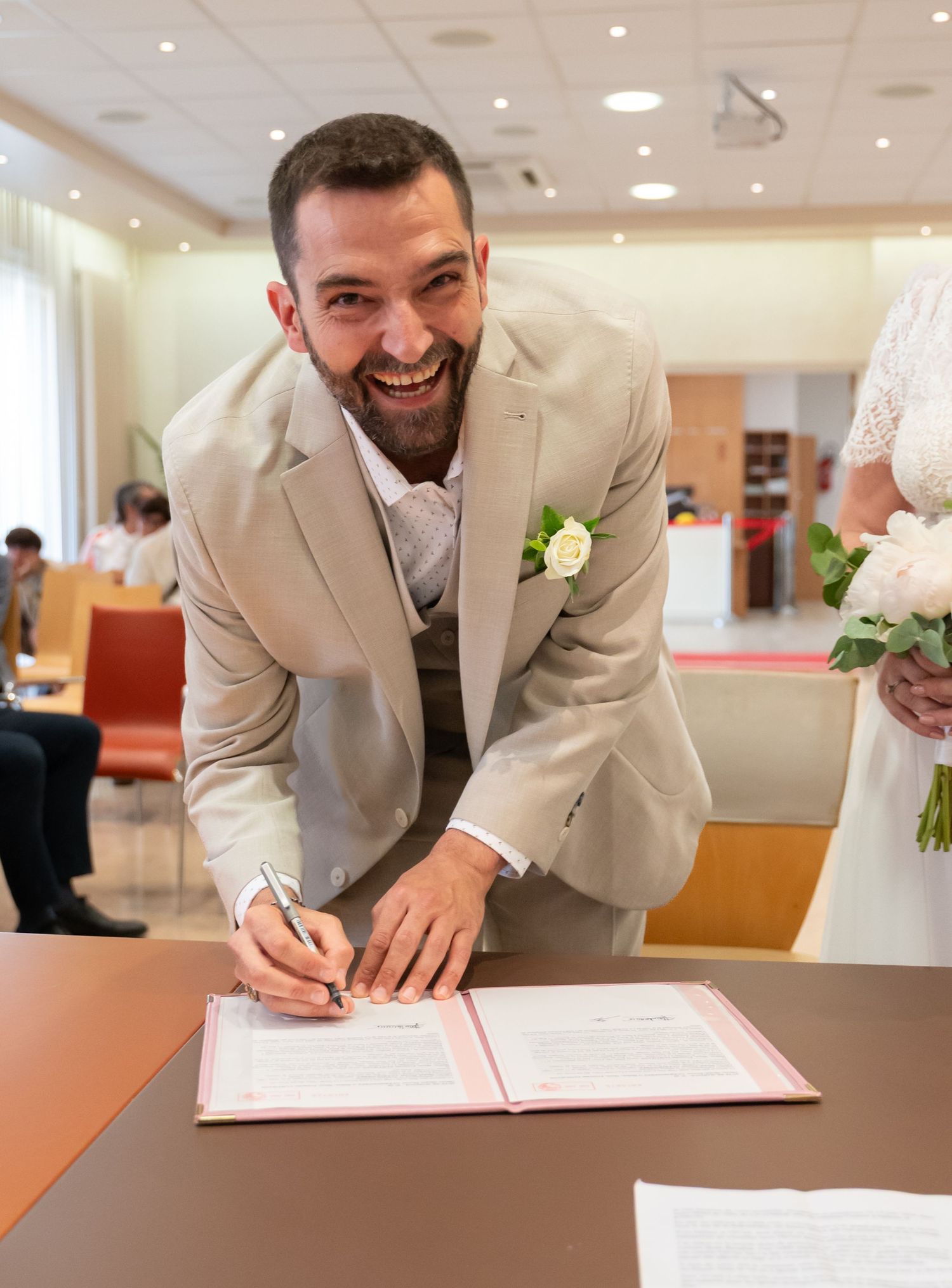 Person in beige suit signing marriage documents with a joyful expression.