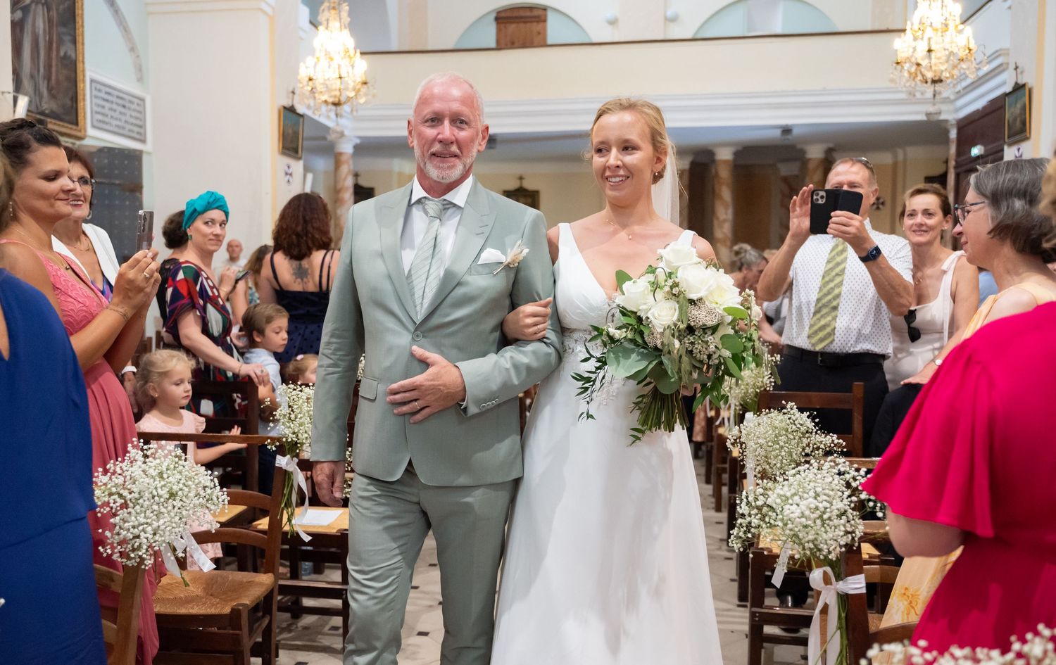 Wedding ceremony in elegant church with chandeliers and guests seated in pews.