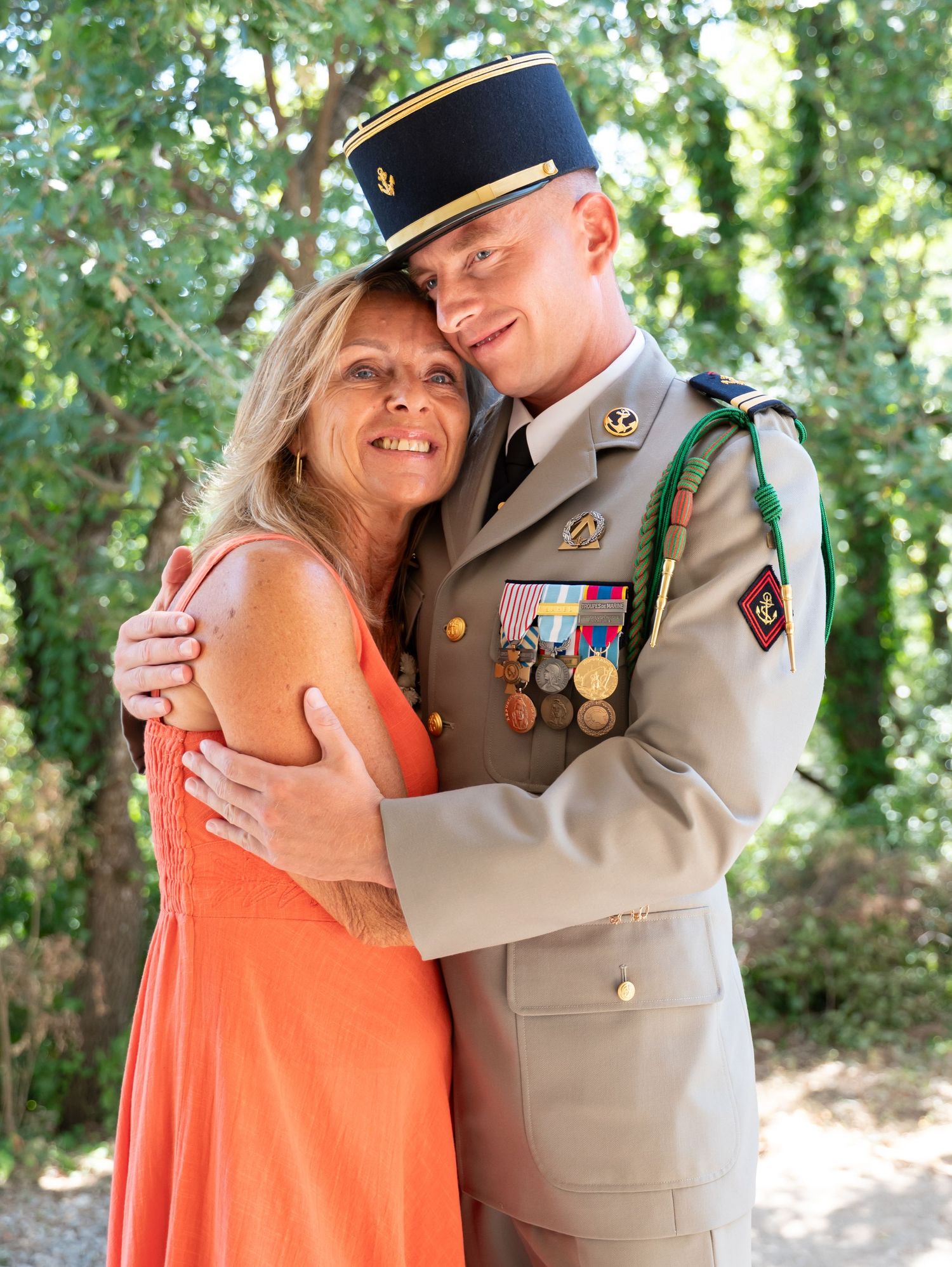 Couple embracing outdoors, with one wearing bright coral dress and other in military dress uniform.