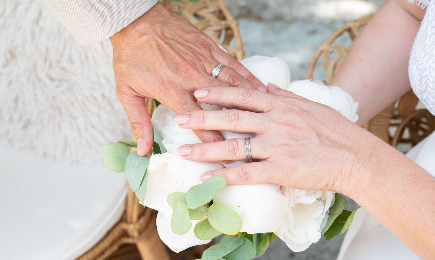 Close-up of hands with wedding rings against white floral bouquet.