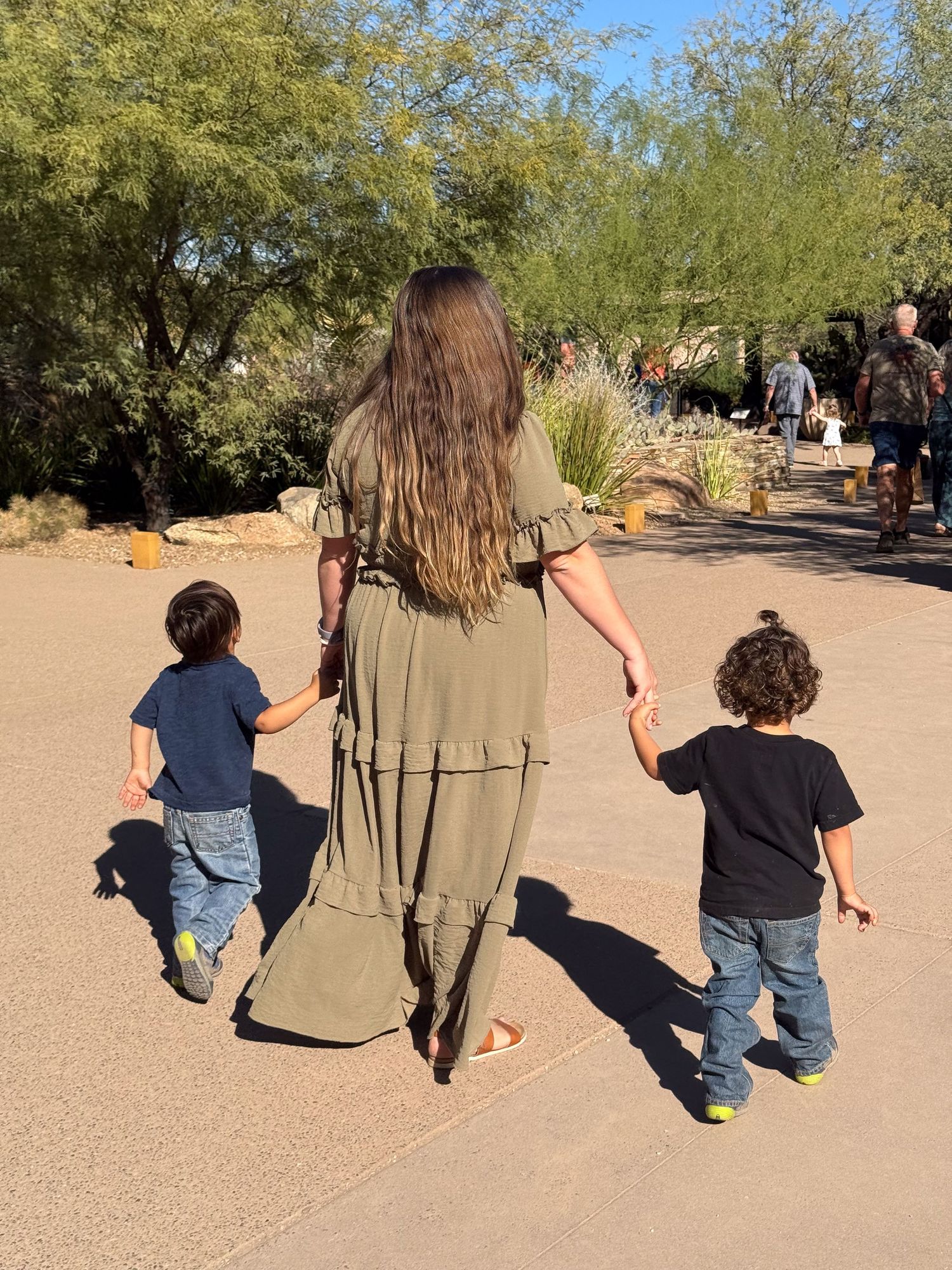 A person in a long green dress holds hands with two small children while walking on a desert path with trees.