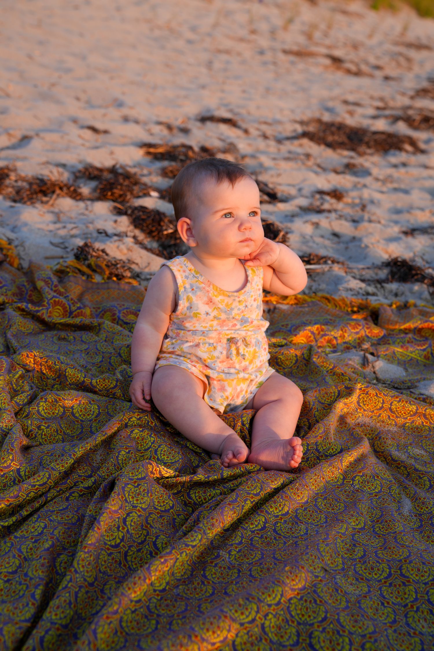 A baby in a floral romper sits on a patterned blanket at the beach during golden hour sunset.