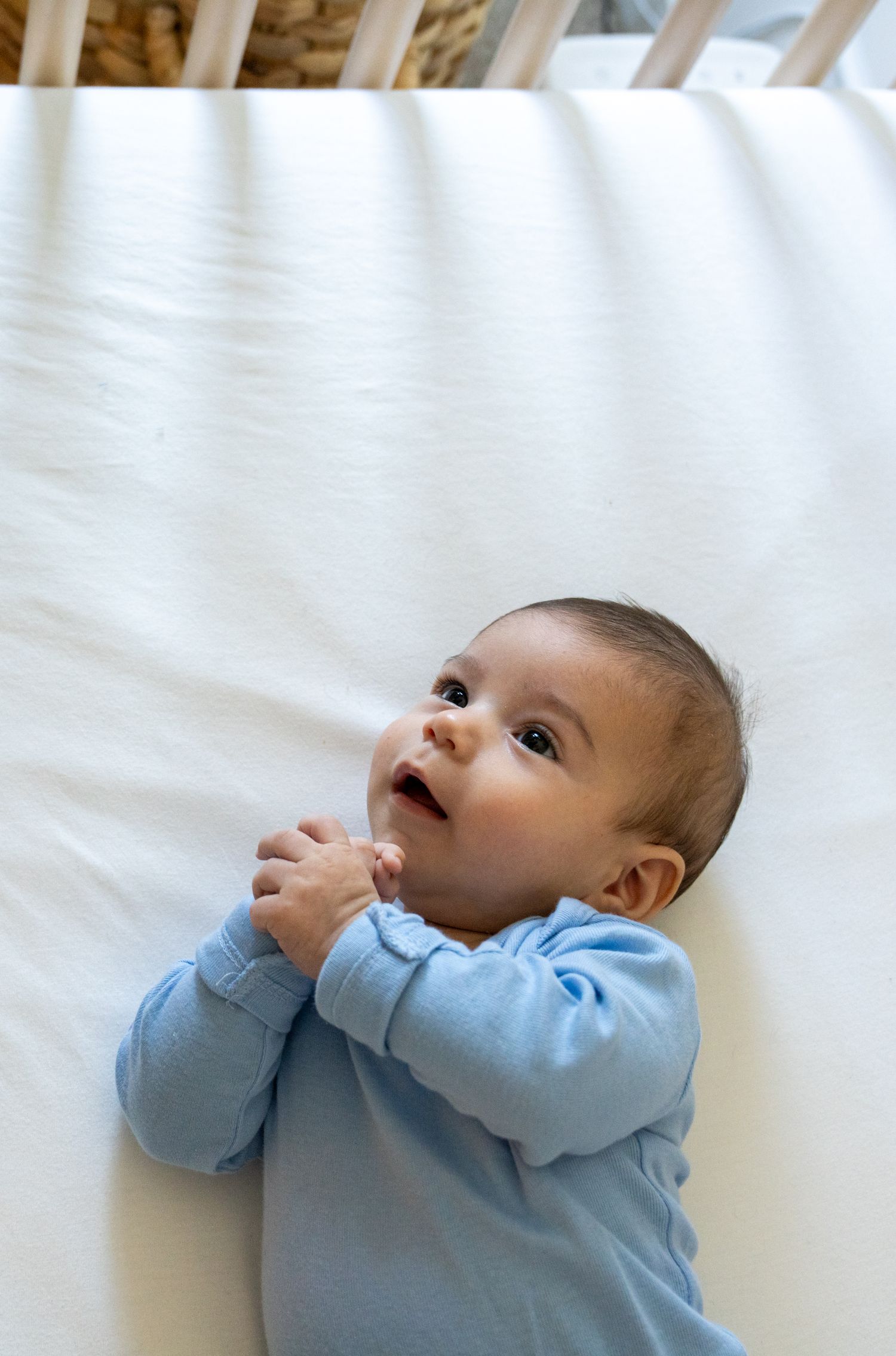 Baby in light blue sweater lying on white bedding looking up with hands near mouth.