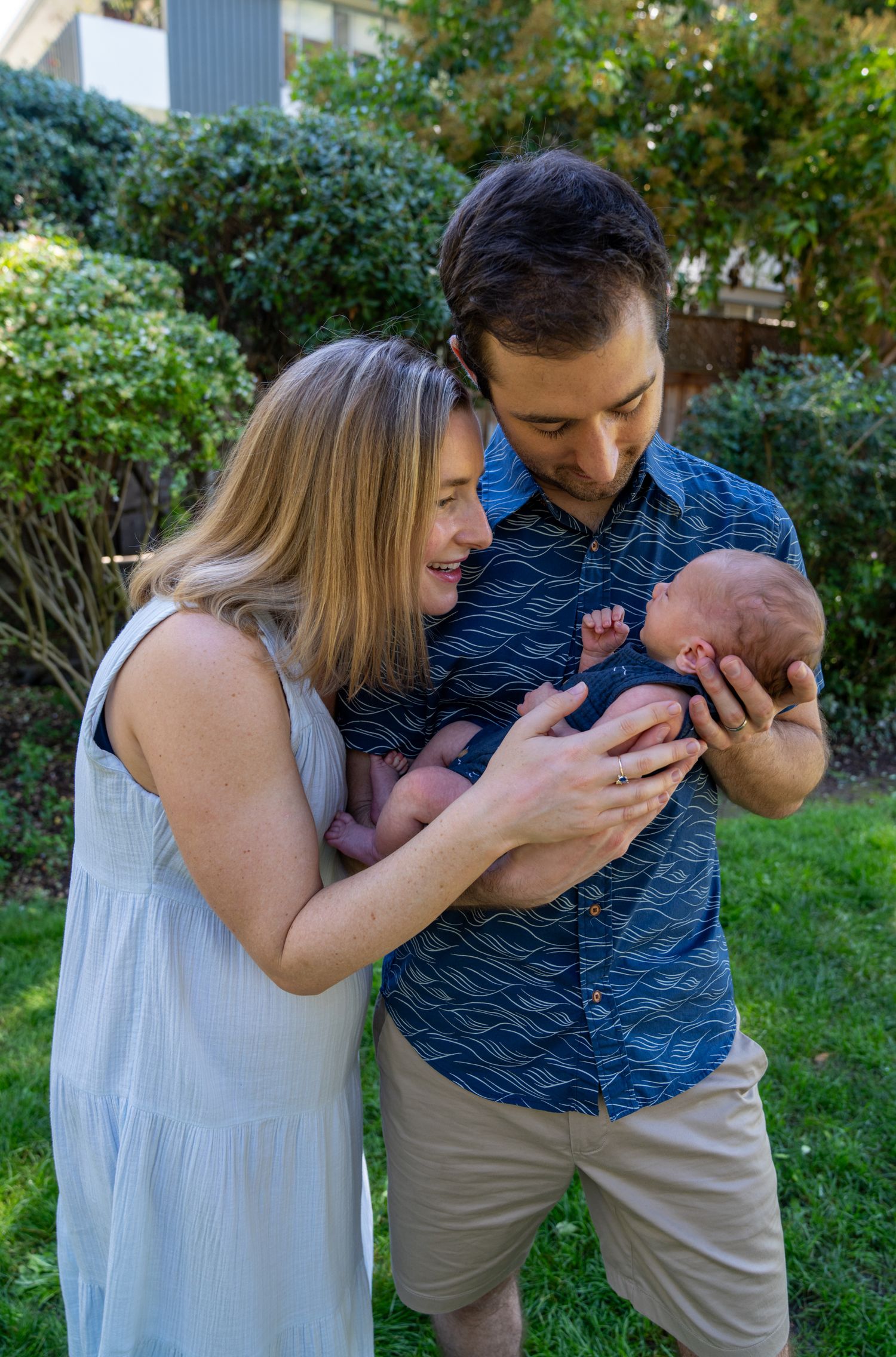 A couple stands in a garden admiring their newborn baby together on a sunny afternoon.