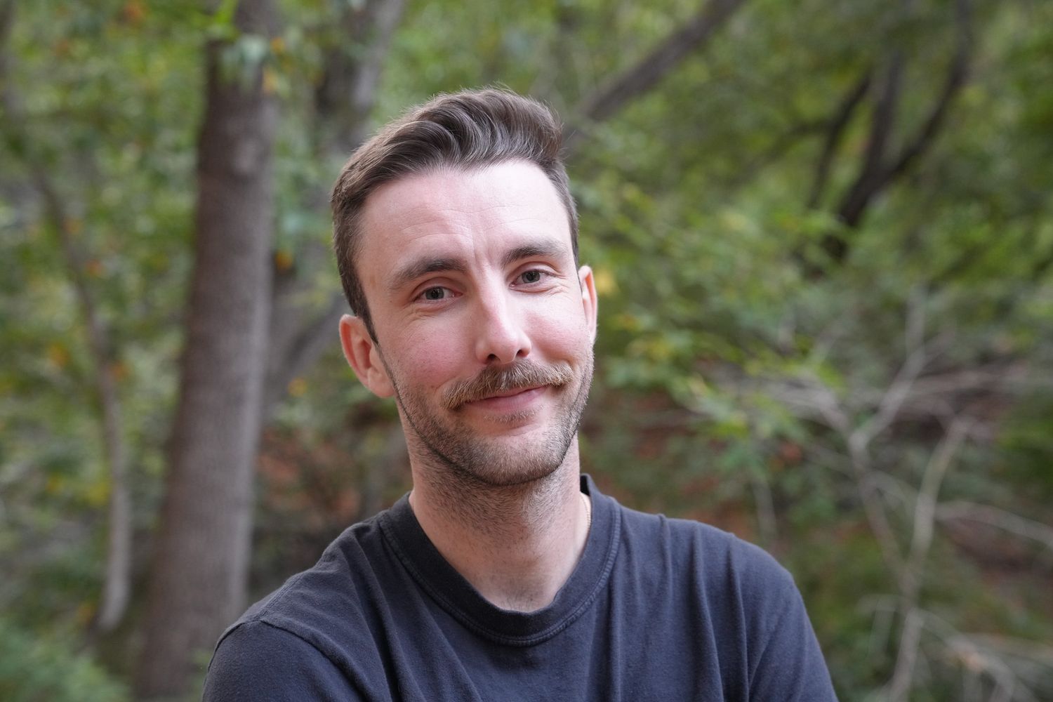 A person with a mustache is shown in a front-facing photo against a blurred outdoor forest background. Young man in black t-shirt