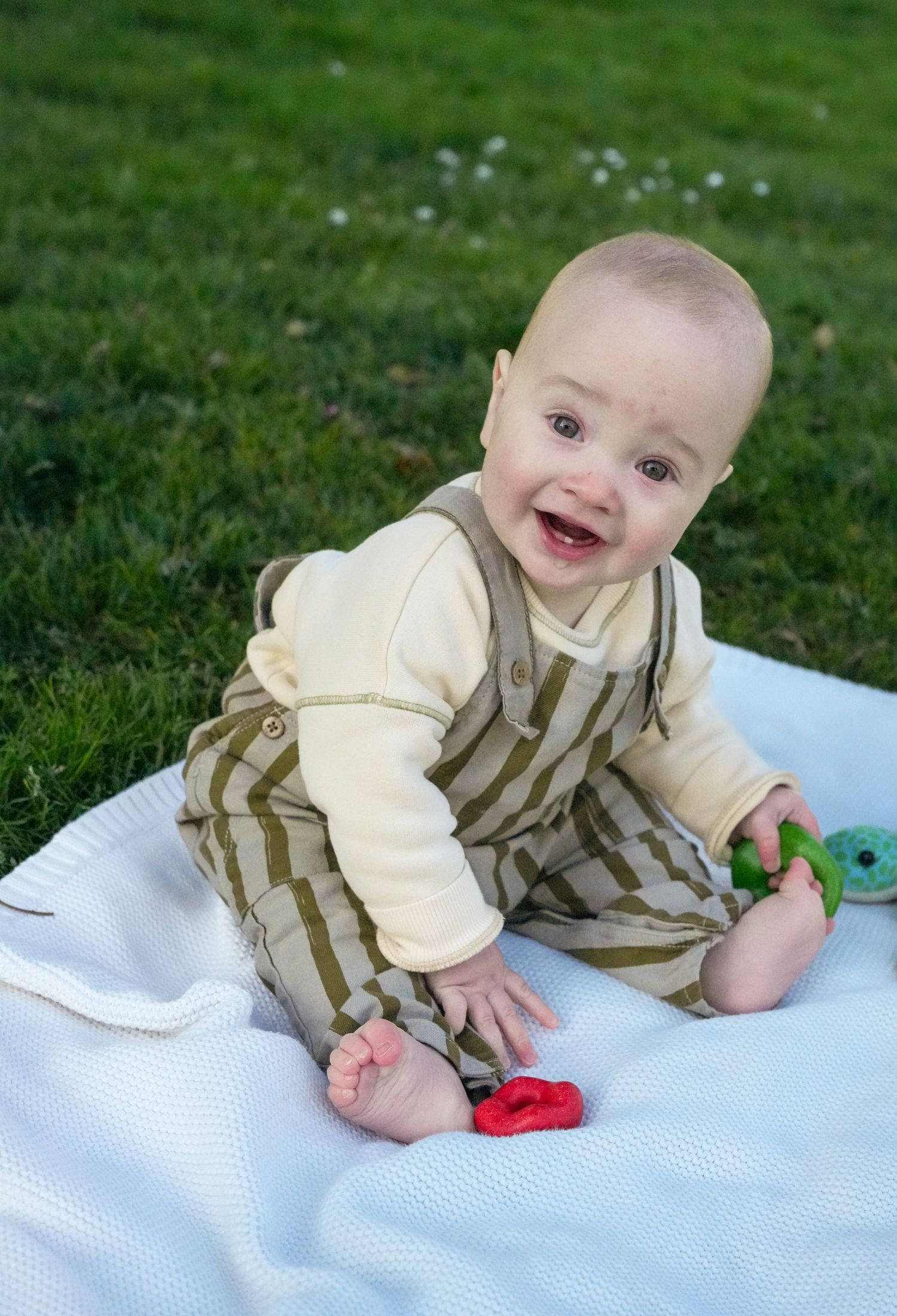 Baby in striped outfit playing with toys on white blanket outdoors.