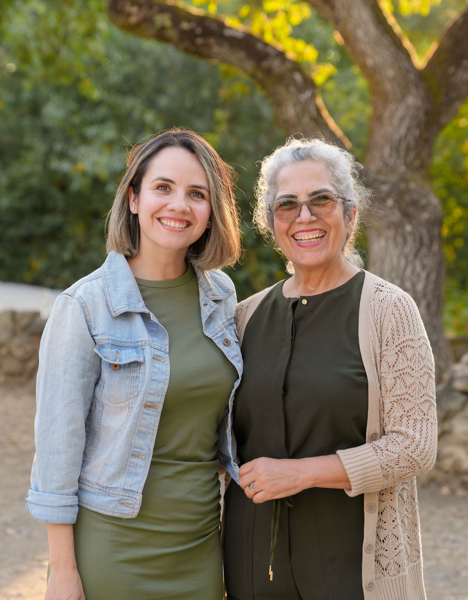 Two people pose together wearing casual outfits against a backdrop of autumn trees.