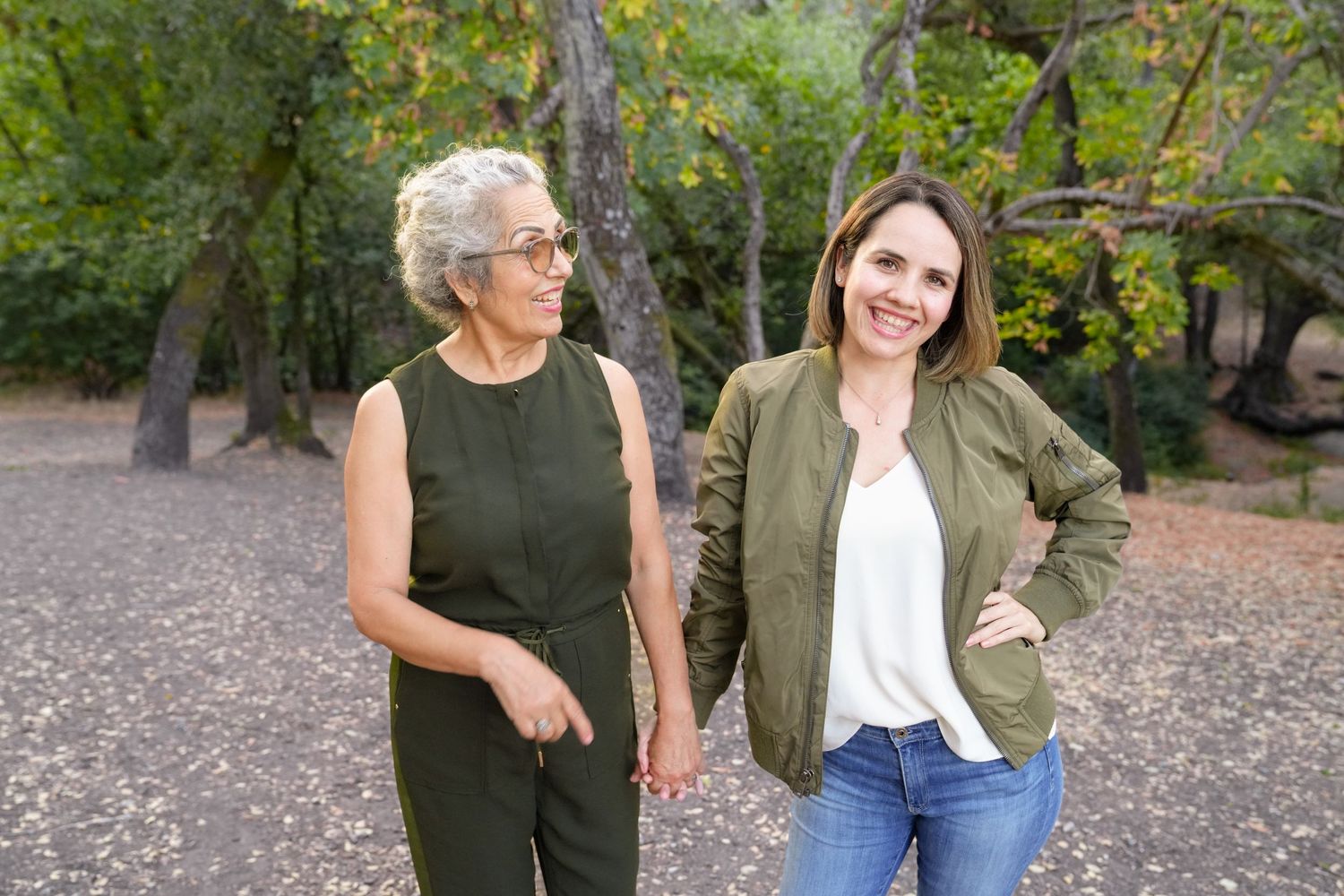 Mother and daughter stand together outdoors in a park with trees and fallen leaves in the background.