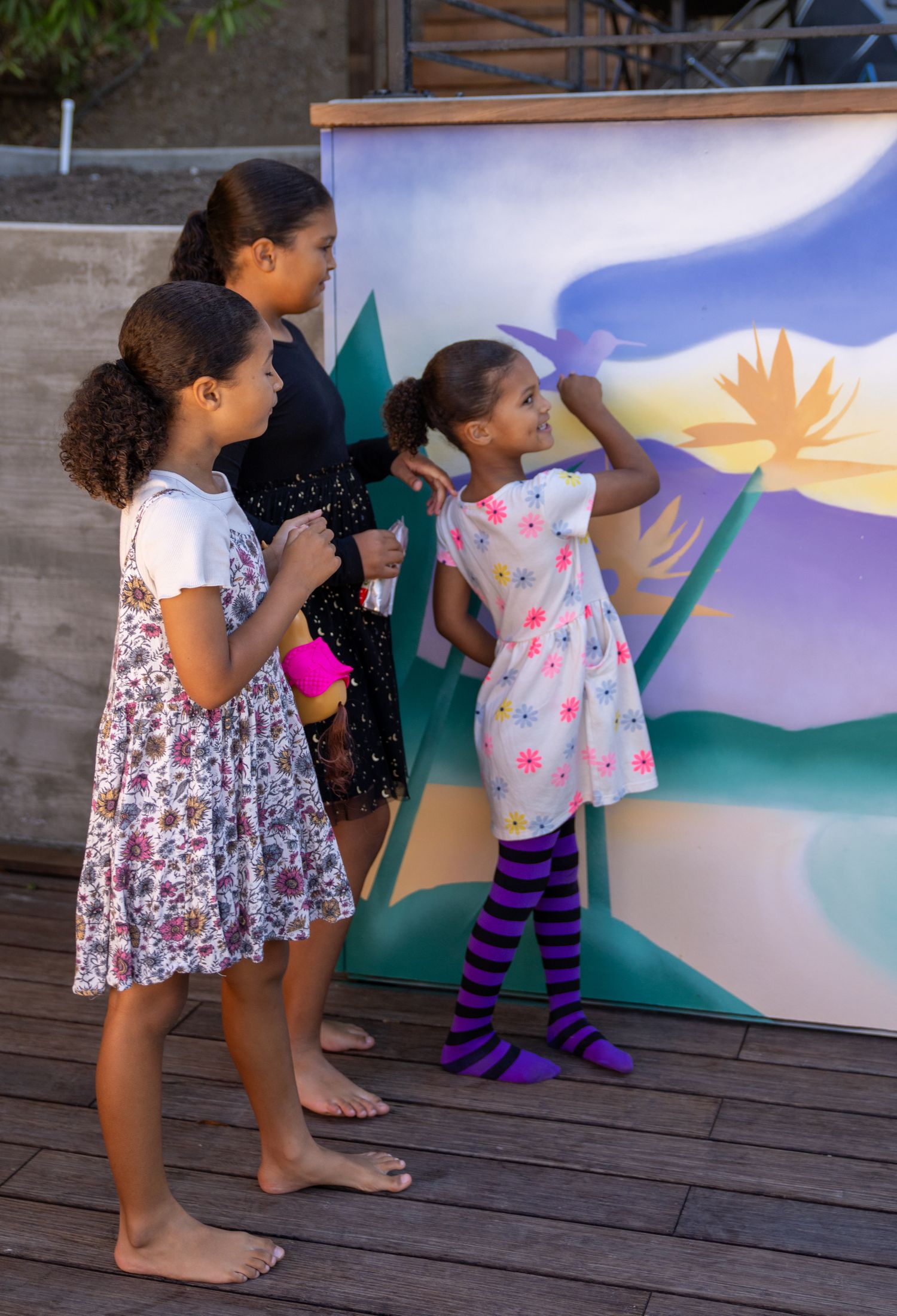 Two young children paint together on a large canvas outdoors, creating colorful artwork on a wooden deck.