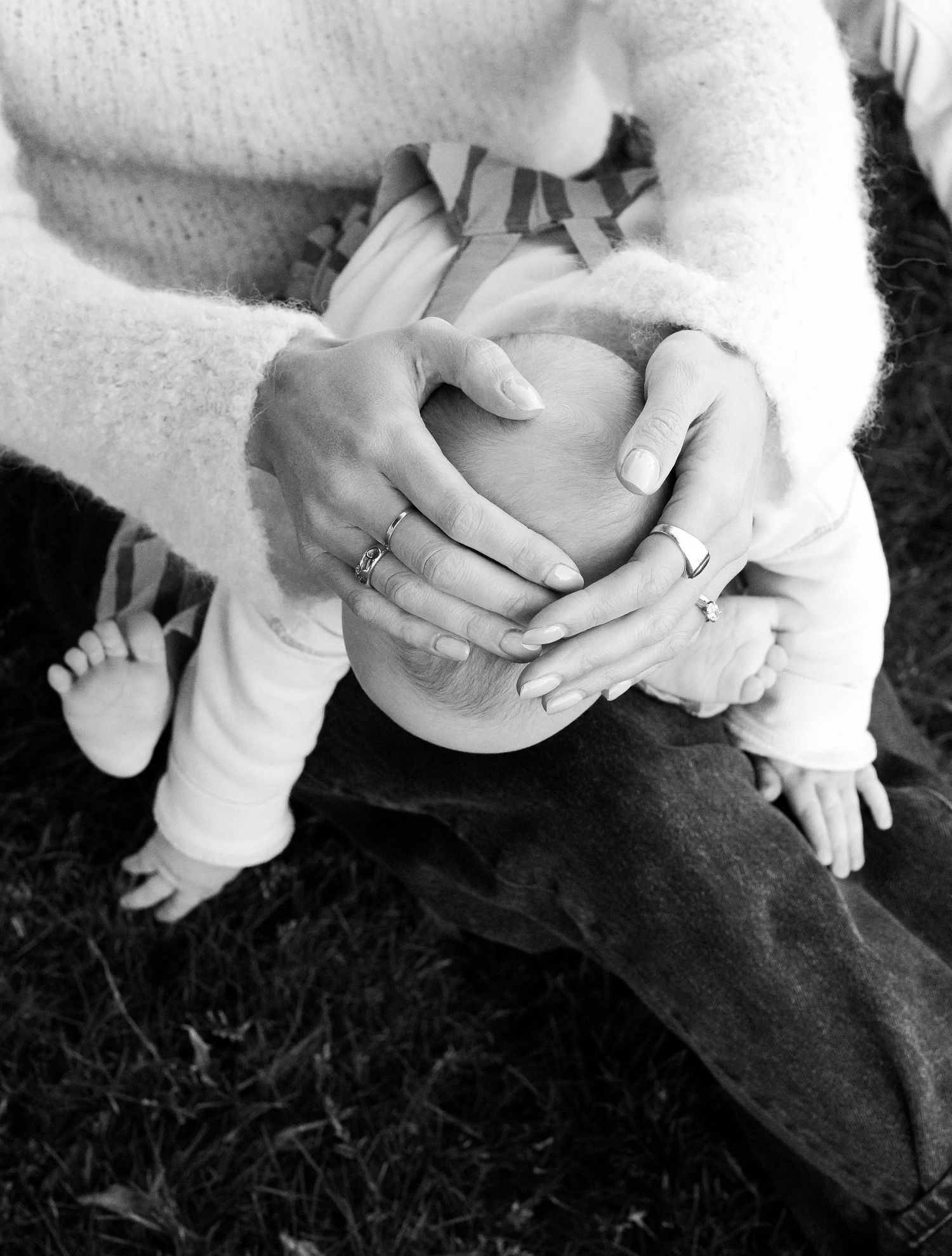 Black and white close-up of hands cradling a small infant outdoors on grass.