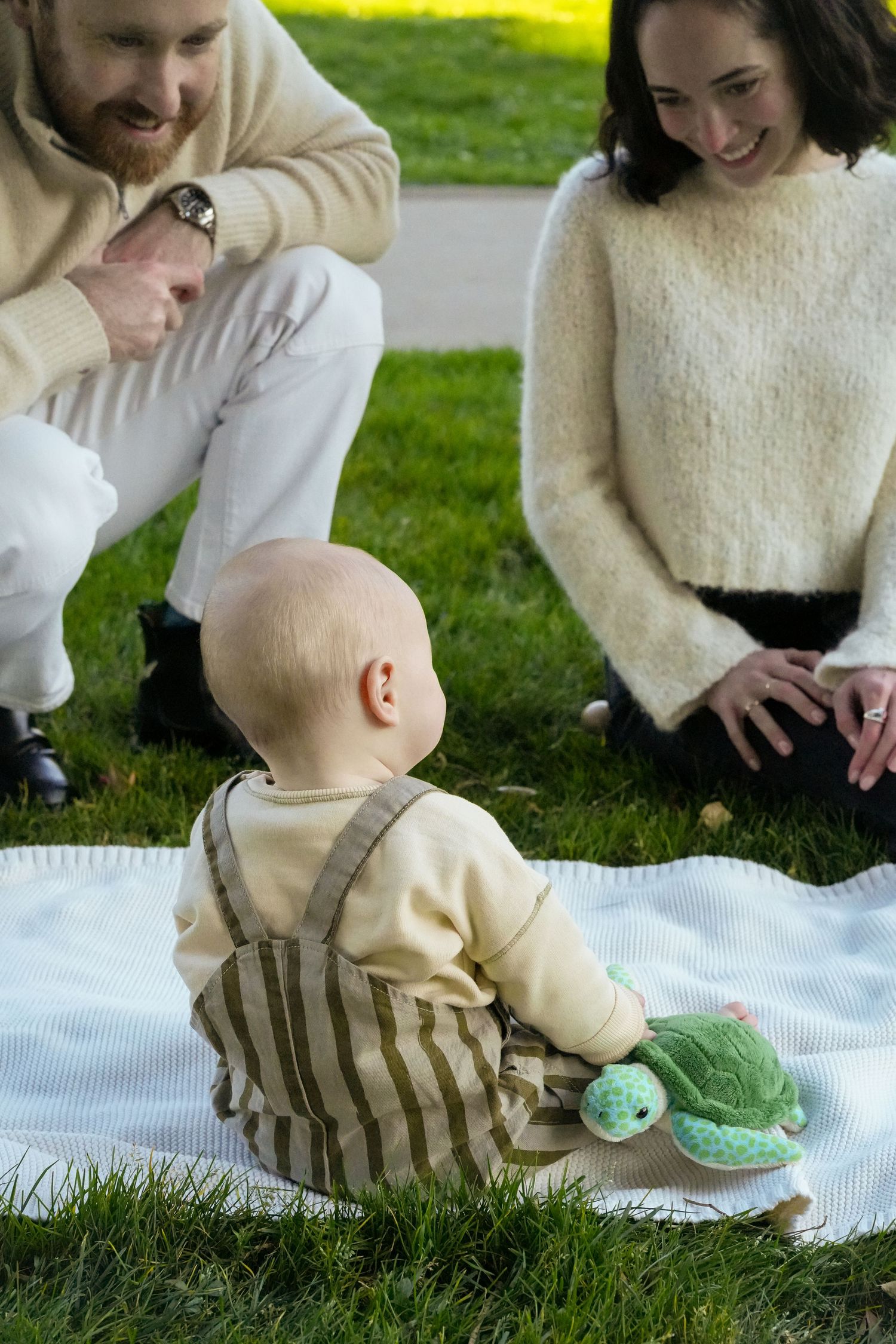 Family sitting together on white blanket in grass with baby playing.