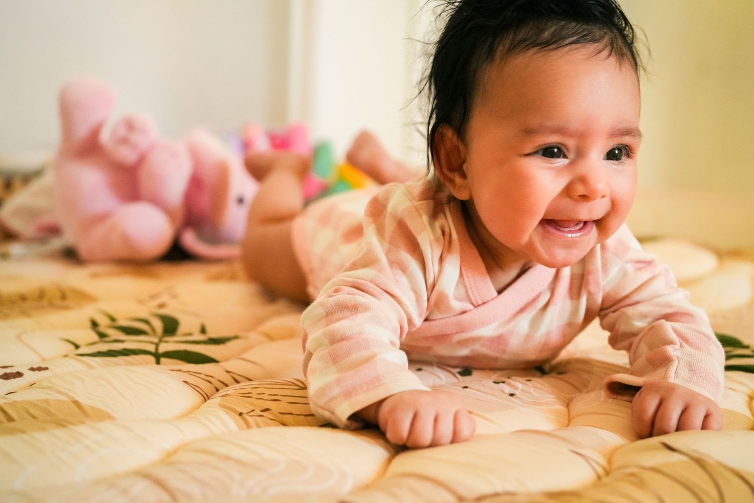 Baby in pink outfit smiling while lying on patterned bedding.