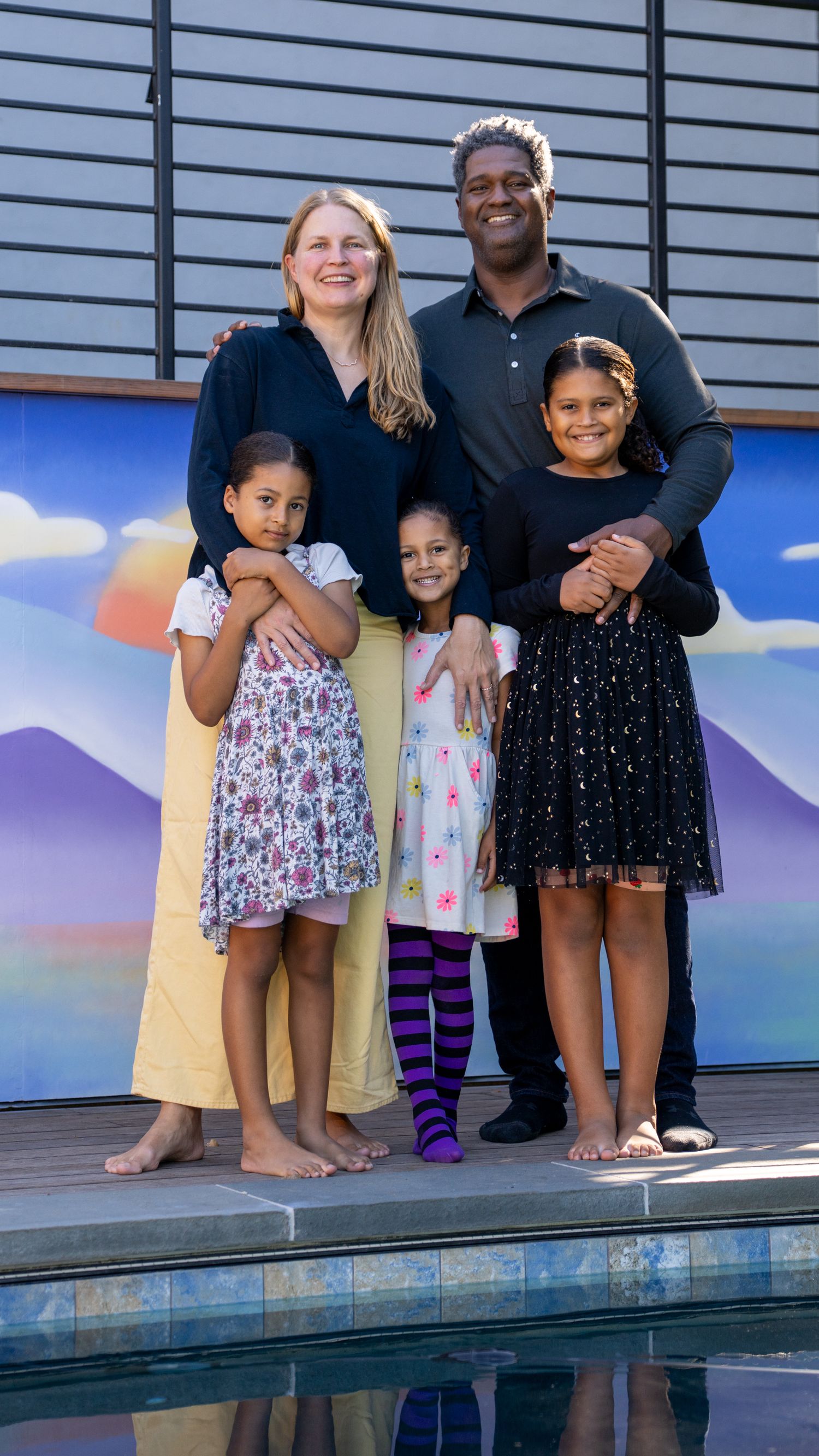Family members stand together at the edge of a pool with a colorful mural backdrop during sunset.