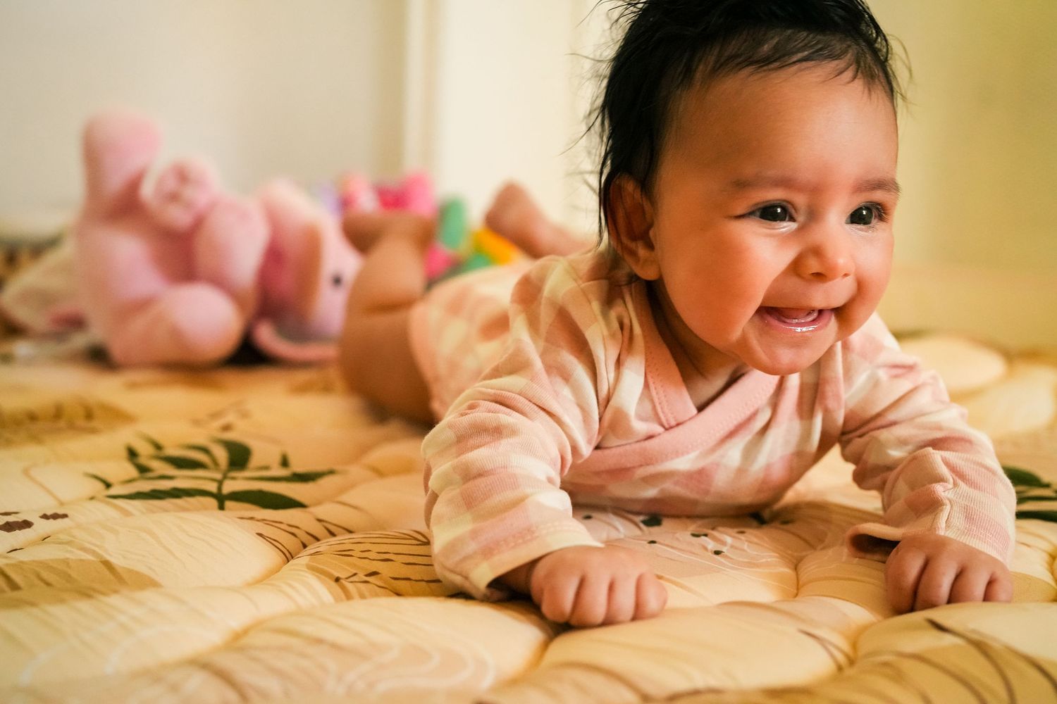 A cheerful baby in pink crawls on a patterned bedspread with stuffed animals visible.