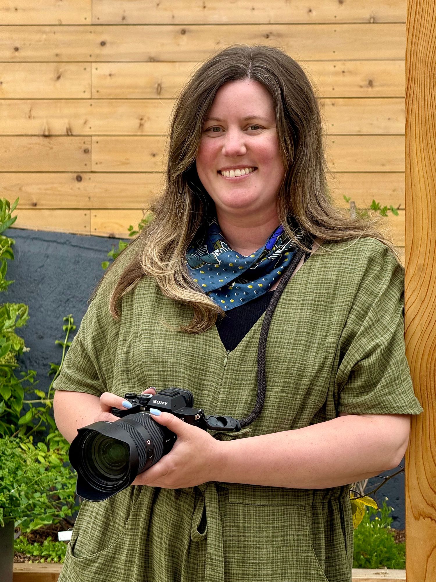 Person in green plaid shirt smiles while posing against wooden paneling outdoors.