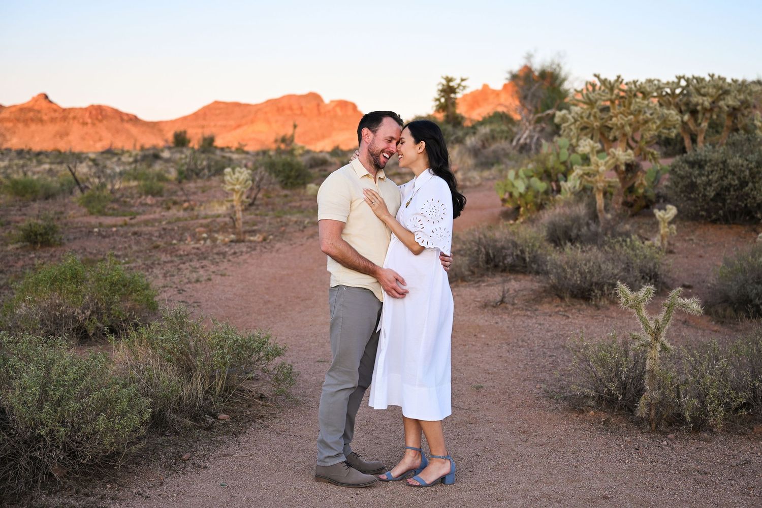 Couple snuggles in the glowing golden hour of the  Arizona Superstition Mountains. Mother is pregnant for their Maternity photos