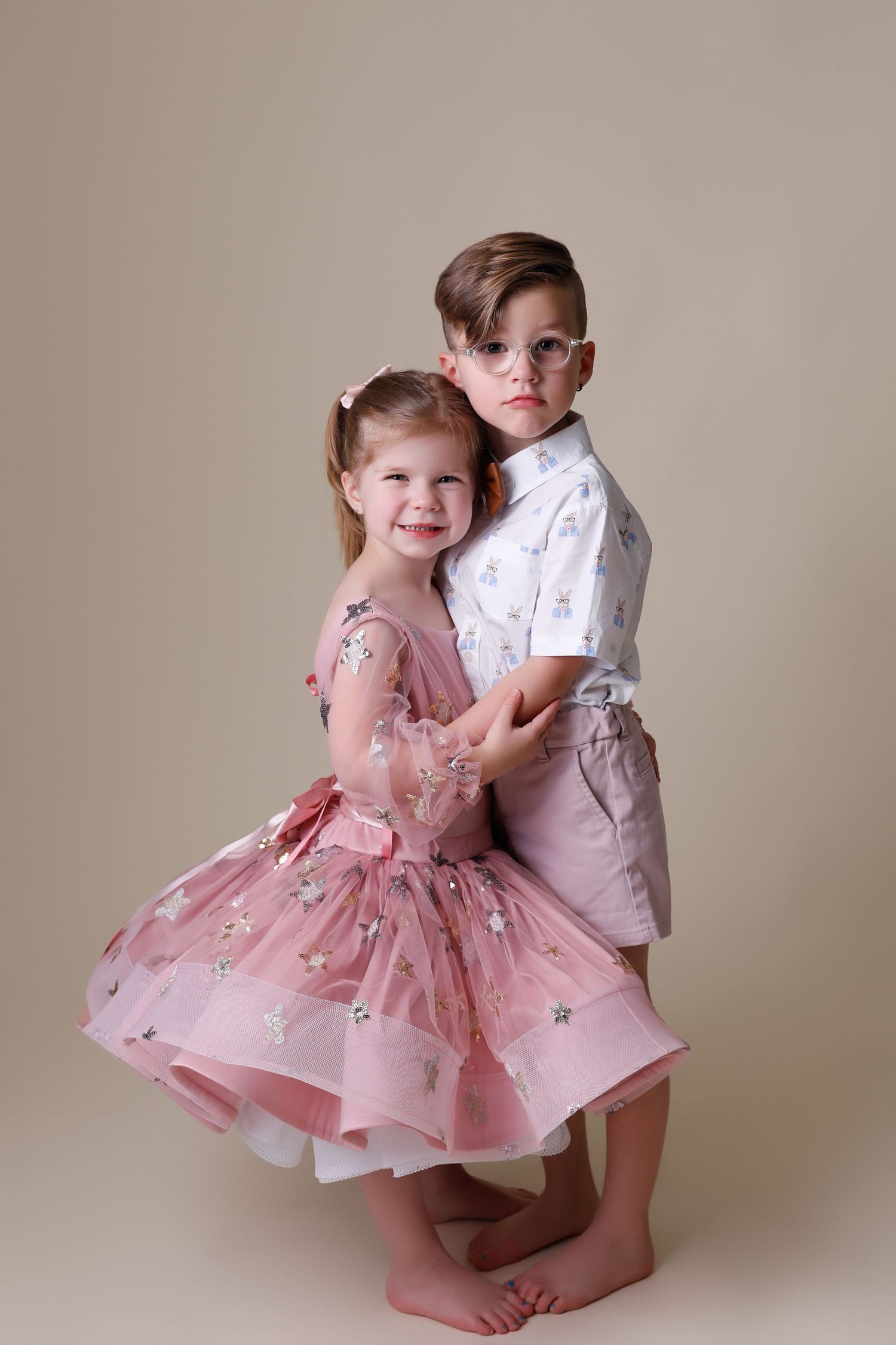 two children stand holding each other on a tan backdrop
