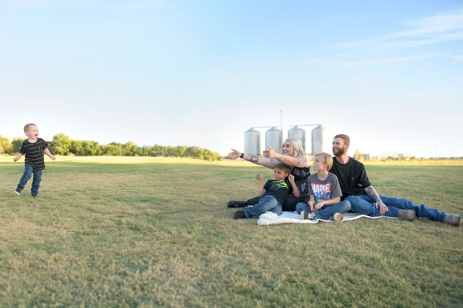 Toddler running towards parents and brothers  in a field of green with silos in the back. Rustic Gilbert Arizona