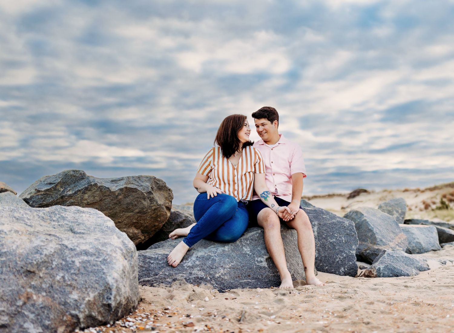 A couple looks lovingly at each other while sitting on a rock with a gorgeous cloudy sky