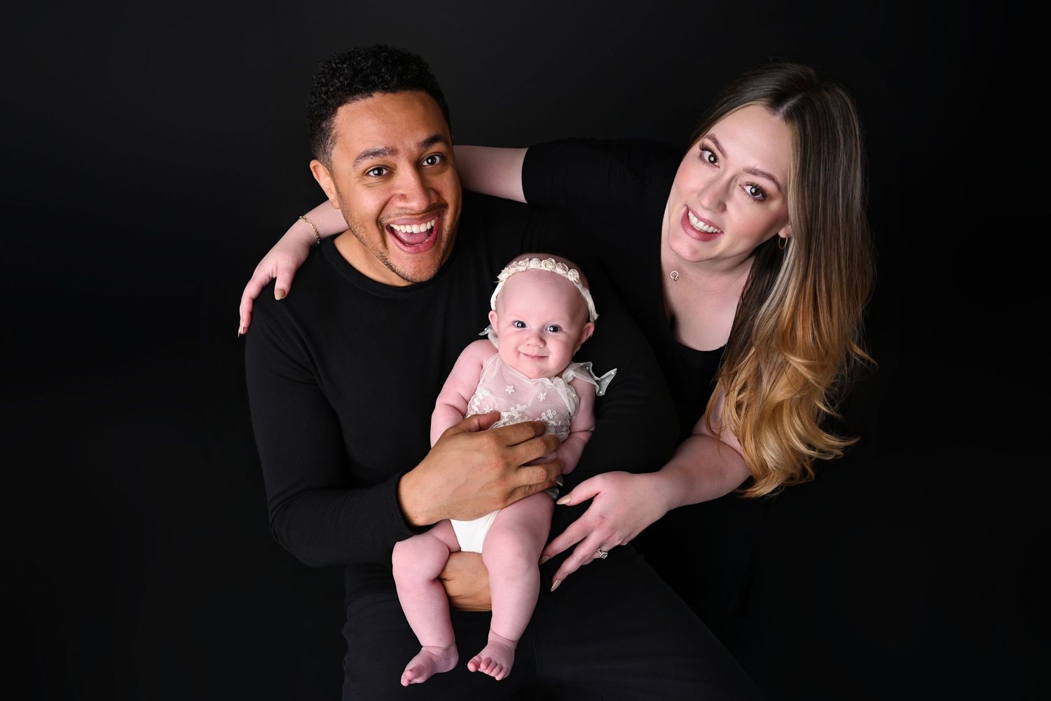 Family of three stand together on black studio backdrop smiling at the camera