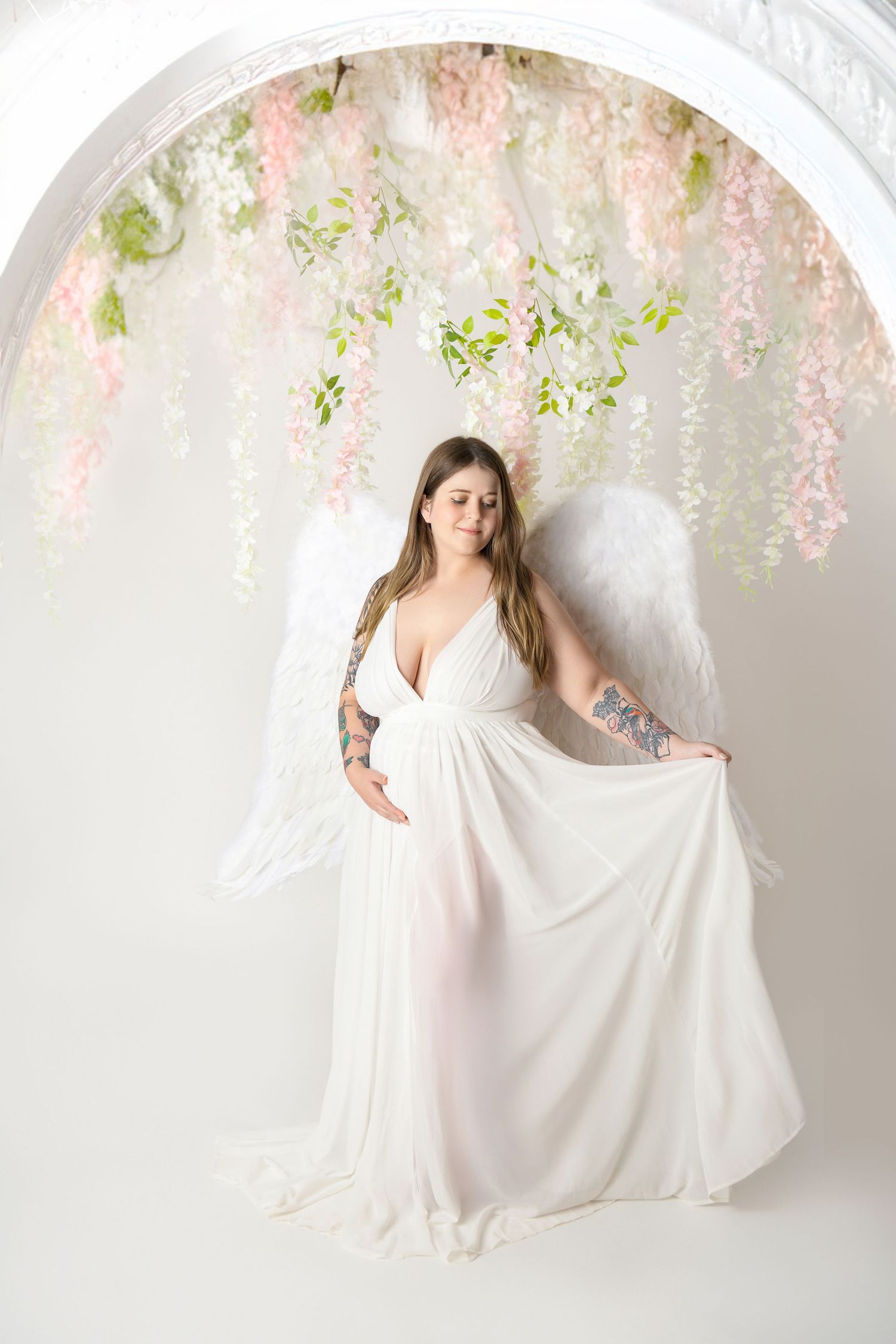 Studio maternity shot with a winged woman gently holding her belly, soft light illuminating her face She is standing under a floral archway
