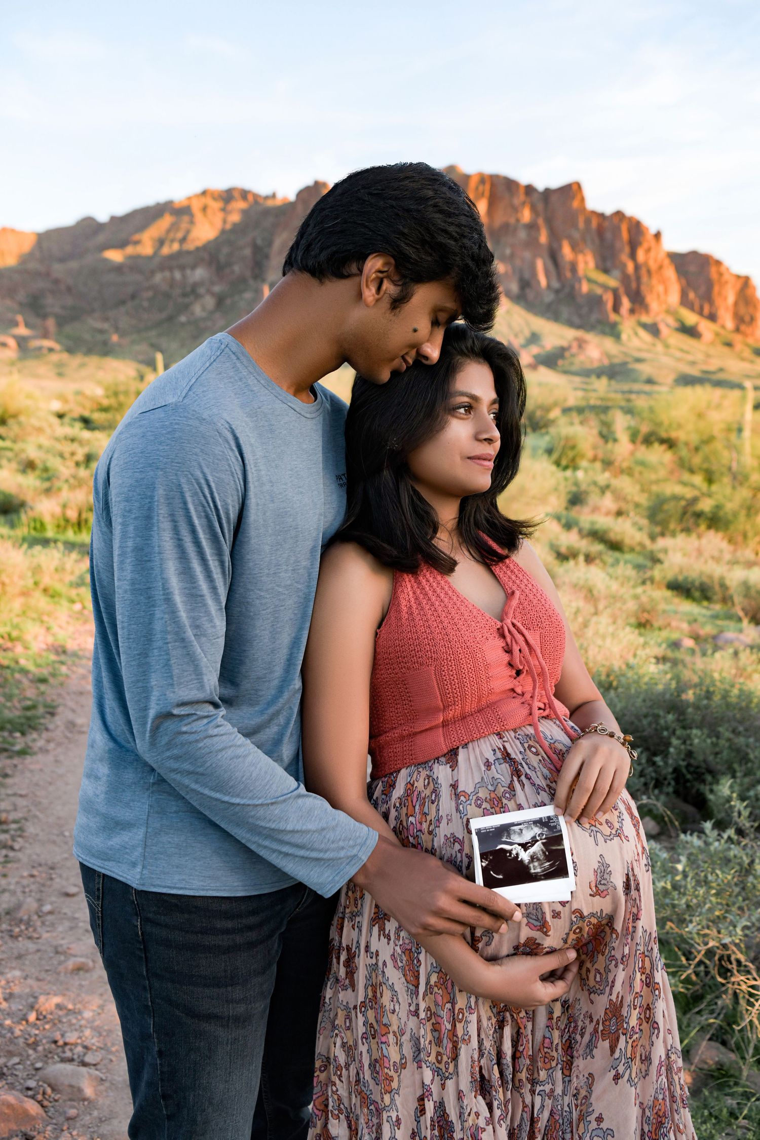 Side profile of a pregnant woman holding her ultrasound photo with her husband in elegant attire with scenic views.