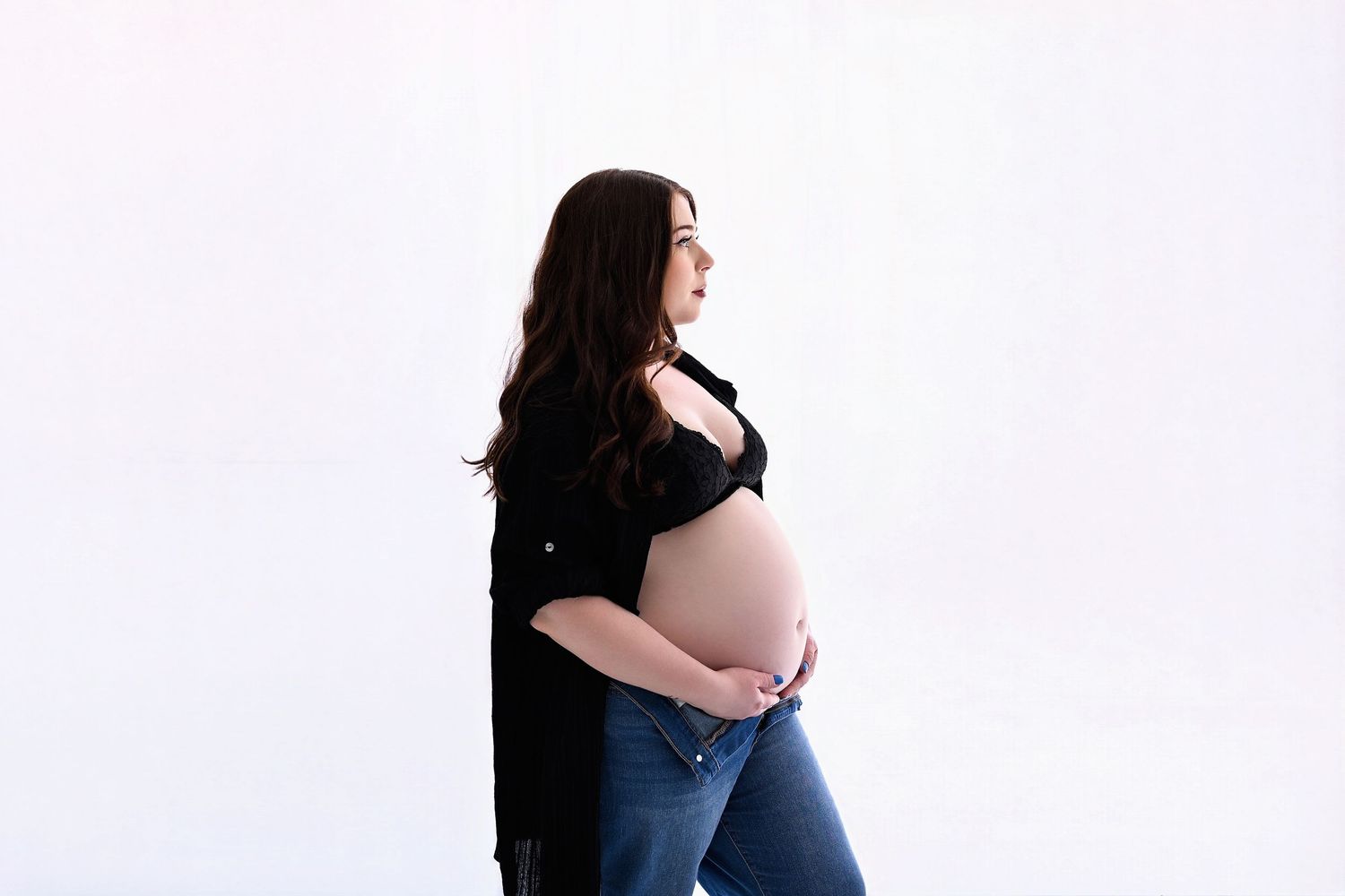 Backlit image of a pregnant mother with her stomach out wearing a lacy bralette and black top that is unbuttoned