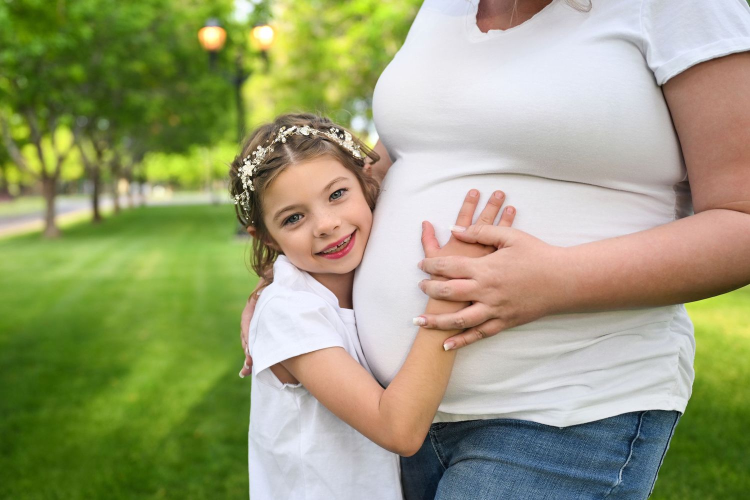 Daughter stands staring into camera while holding her Mothers pregnant belly in an all white gown in an Arizona park