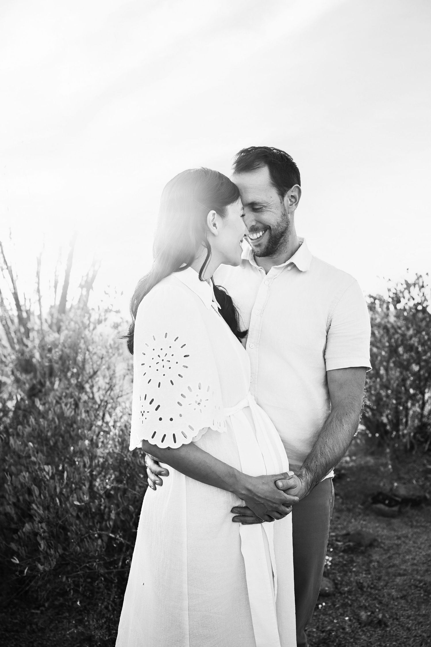 Couple smiling and holding her pregnant belly in a beautiful outdoor setting.